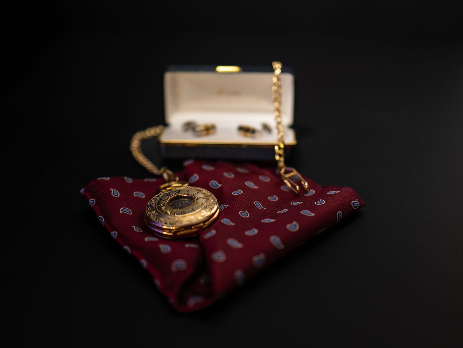 Gold locket pocket watch on burgundy paisley pocket square with cufflinks box in background.