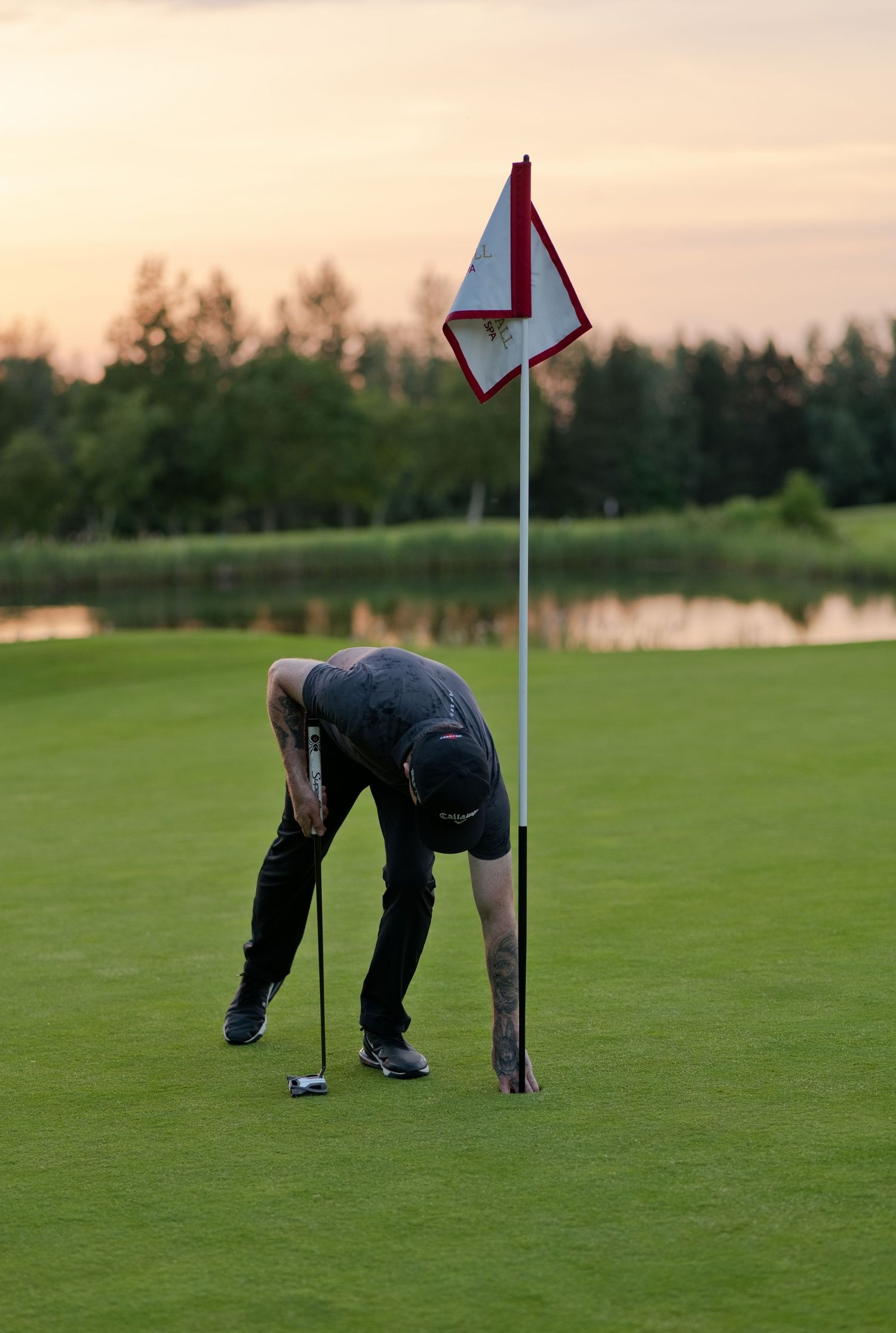 A tattooed golfer bends down near the flag pin on a golf course green at sunset.