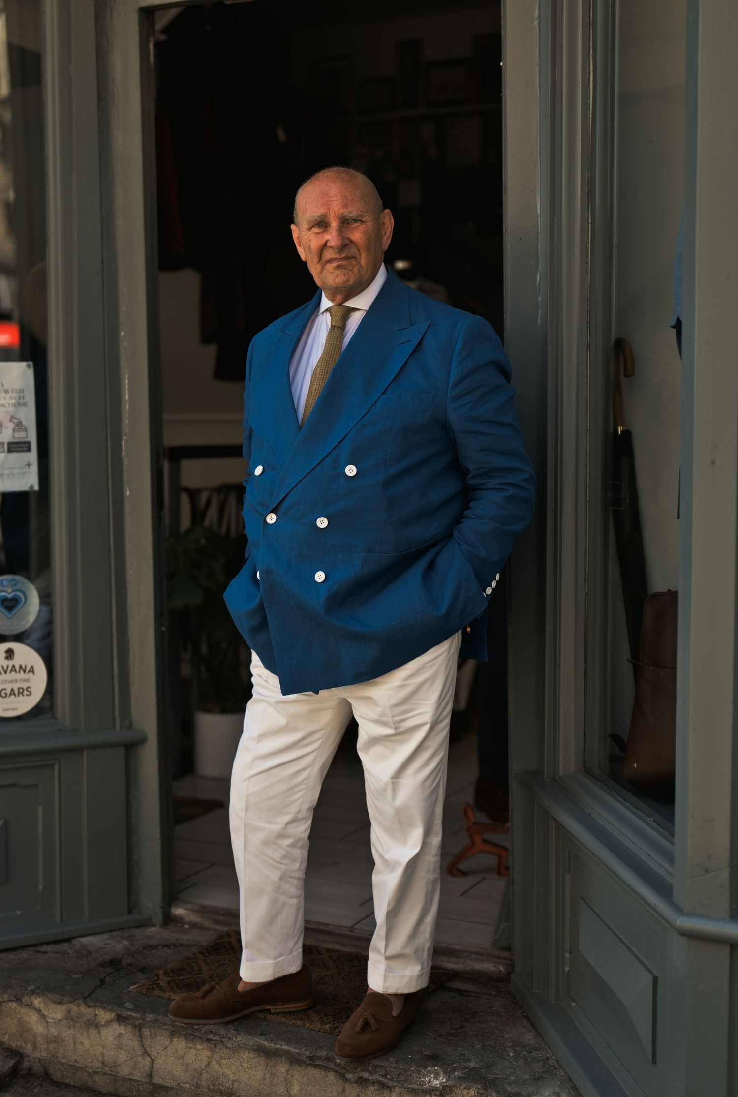 Elegant older man in a blue double-breasted blazer and white trousers standing in a shop doorway.