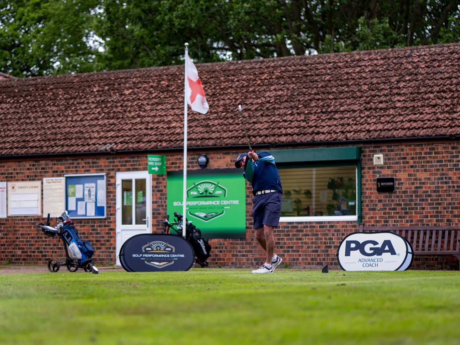 Golfer swings at a PGA golf event with branded signage and brick clubhouse in background.