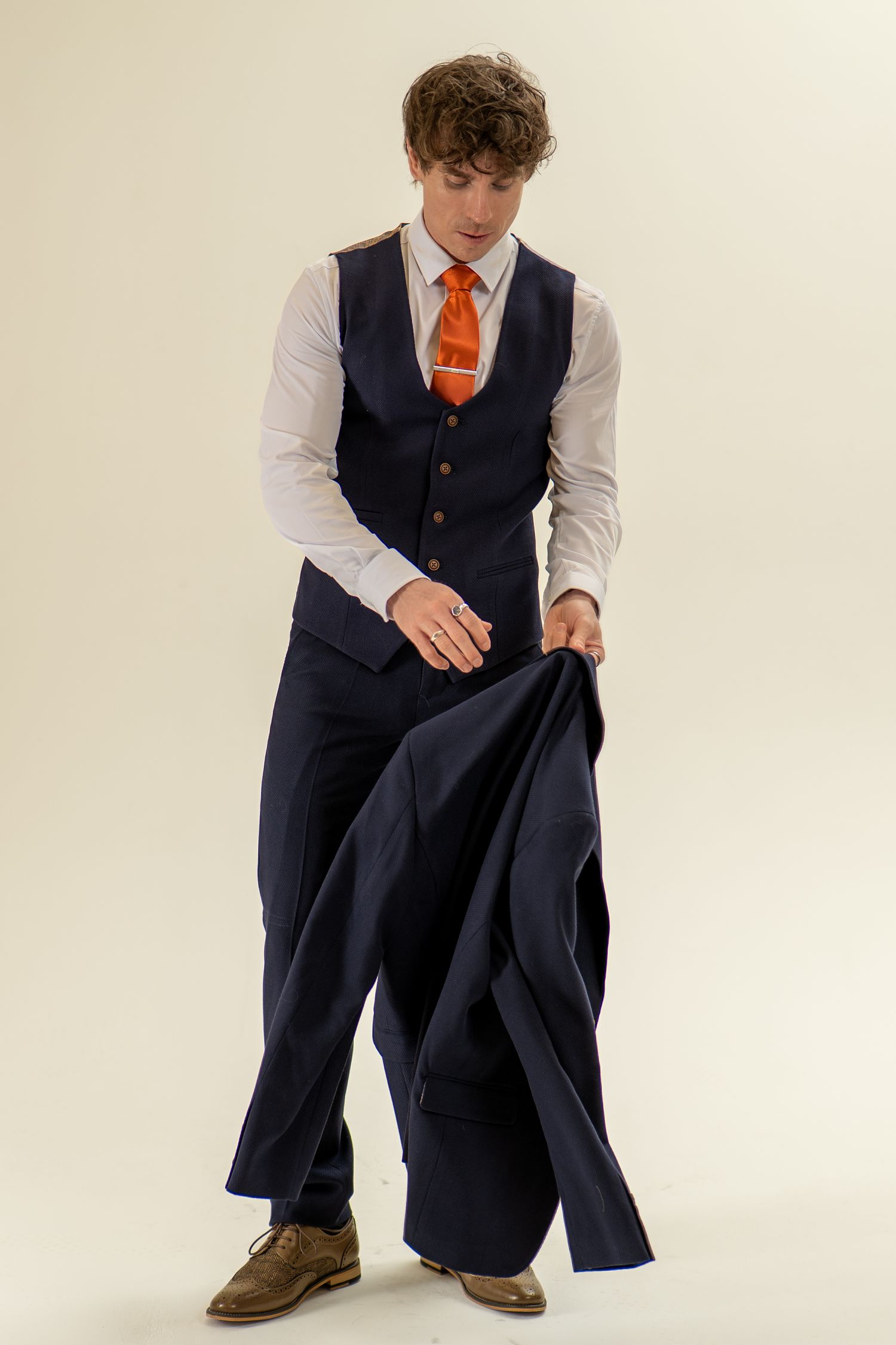 Young man in navy vest and orange tie holding a suit jacket low, studio shot.