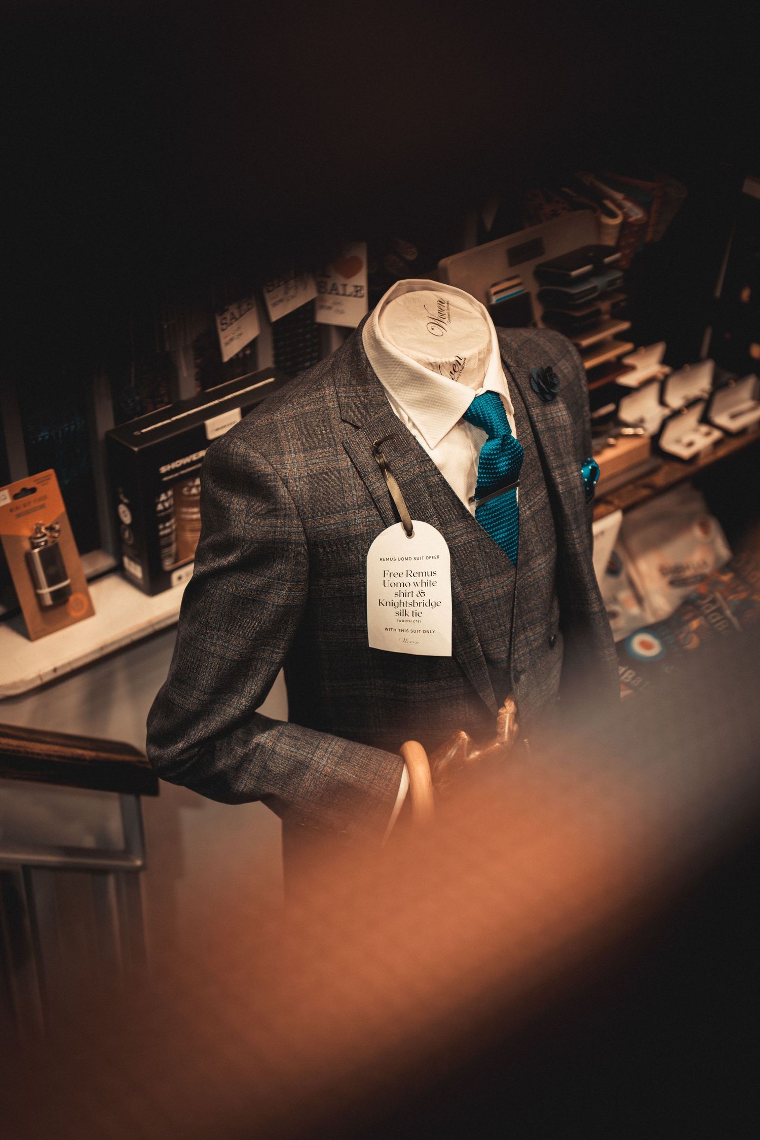 A mannequin displays a grey plaid three-piece suit with a teal tie and price tag in a menswear store.