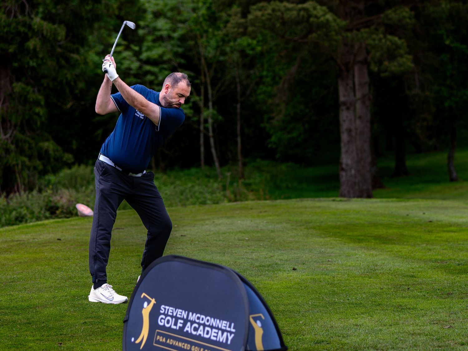 Golfer swinging a club beside a Steven McDonnell Golf Academy banner on a course.