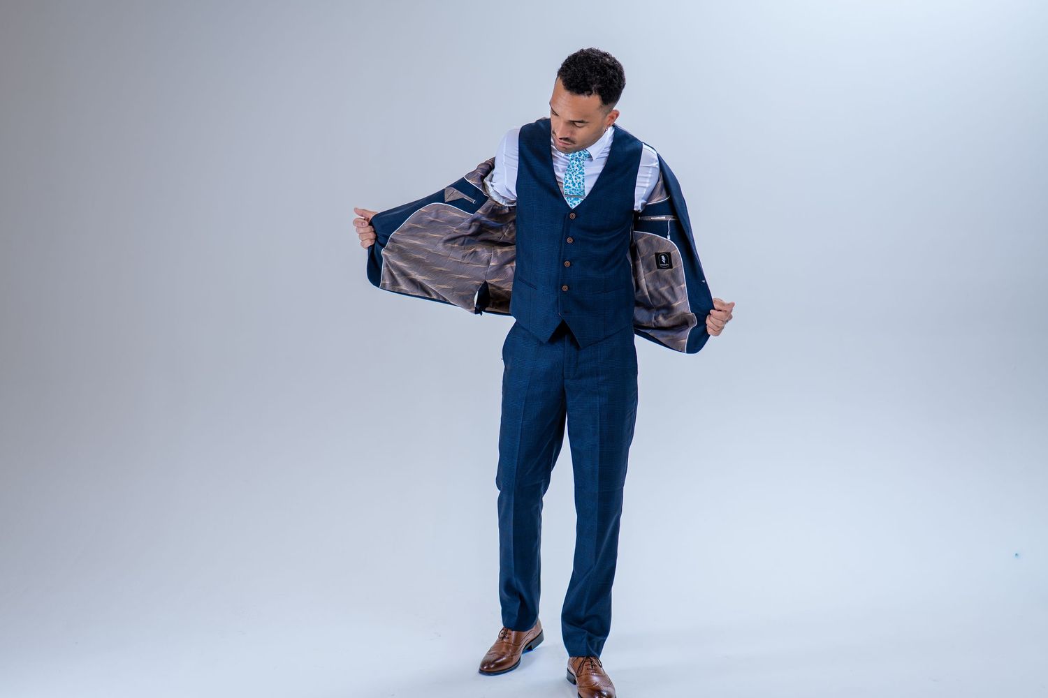 Man in navy blue three-piece suit opening jacket to reveal grey lining in professional studio shot.