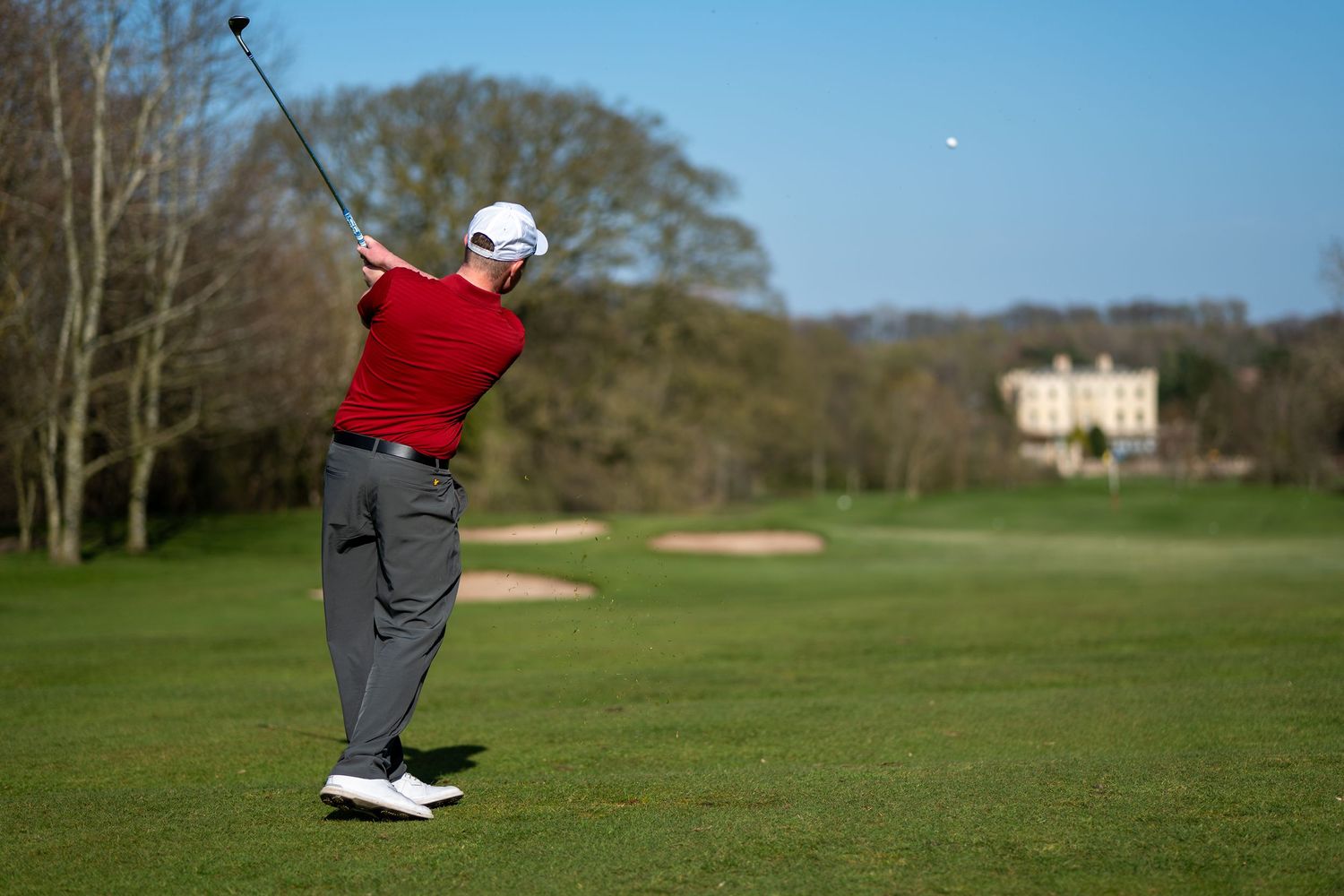 Golfer in red shirt completing a full swing drive on a green golf course fairway.