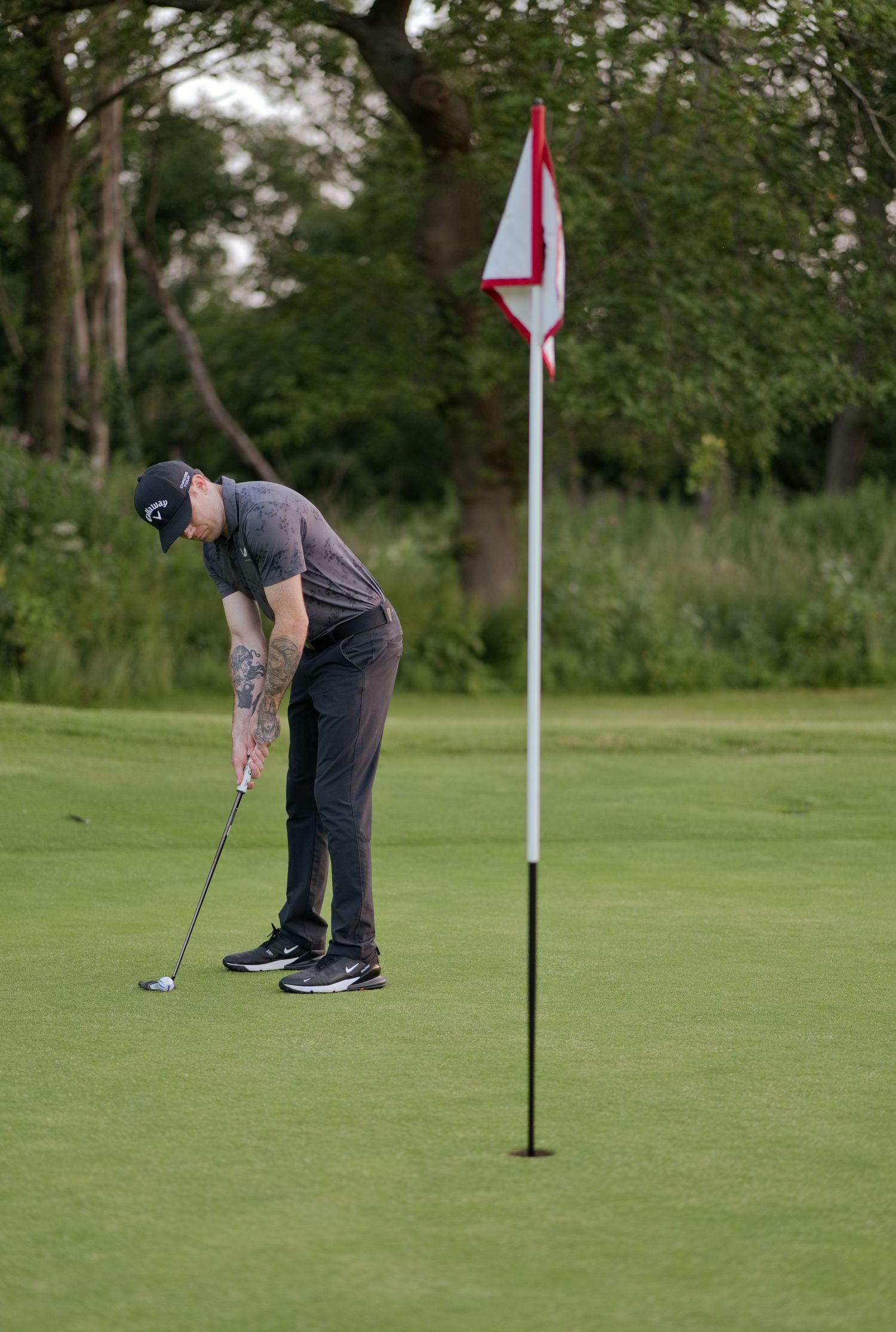 A tattooed golfer in dark clothing putts near a red and white flag on a green golf course.