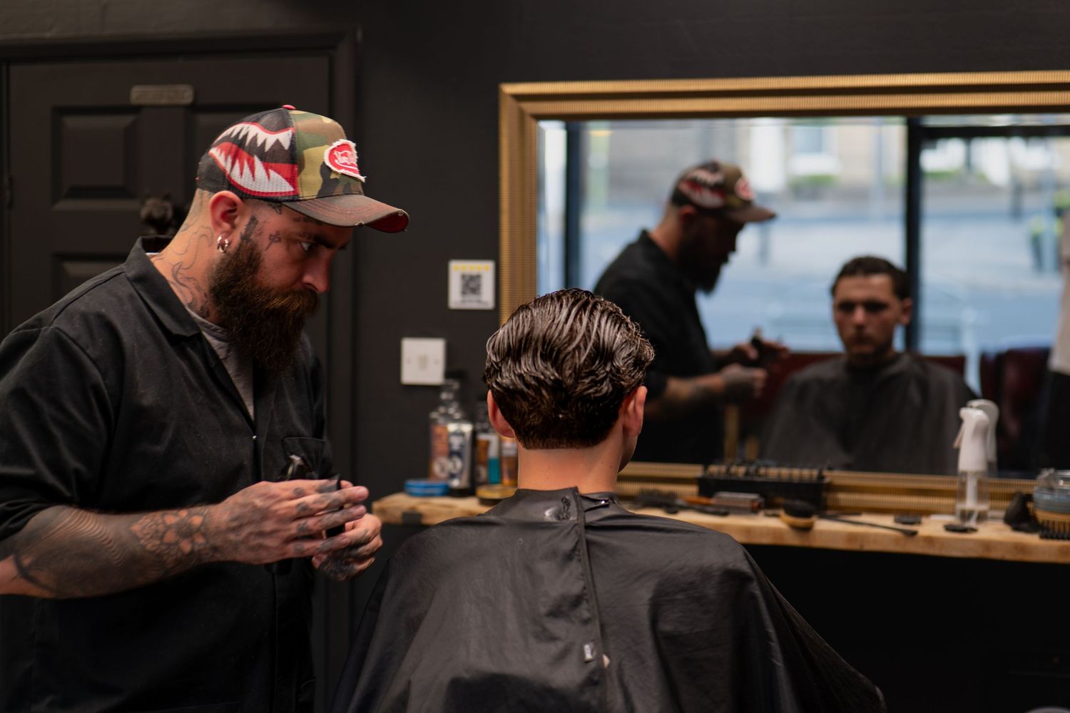 Tattooed barber in camo cap styling client's hair in modern barbershop with mirror reflection.