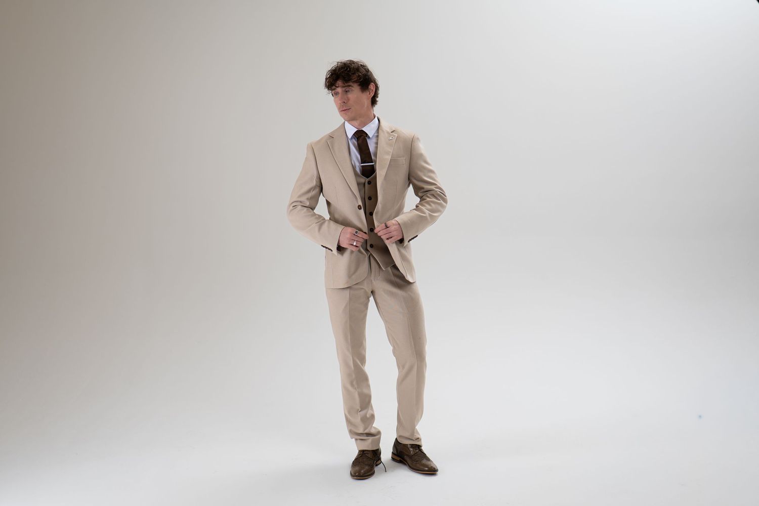 Man in beige three-piece suit buttoning jacket, full-length studio fashion shot.