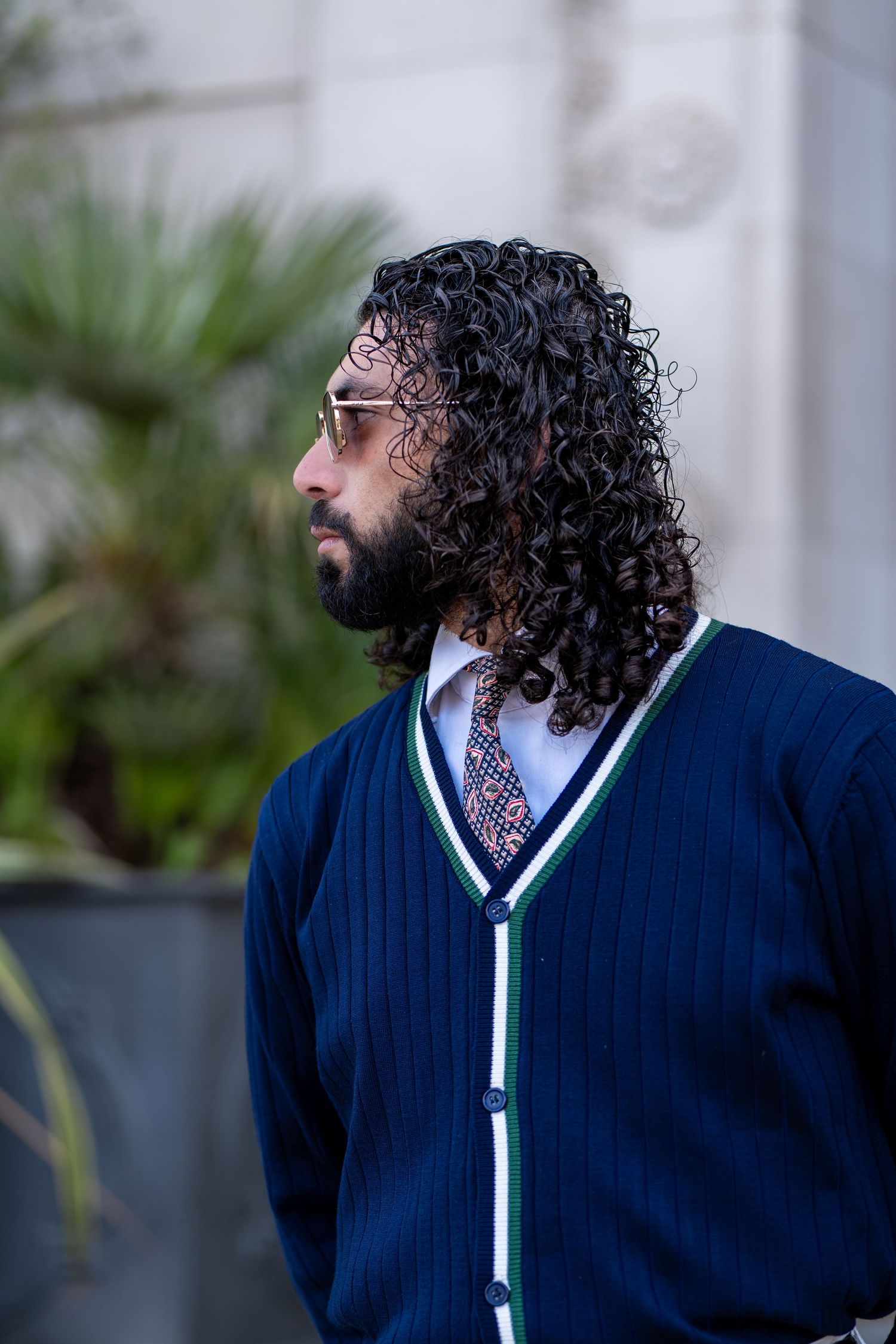 Close-up of bearded man with long curly hair wearing navy V-neck cardigan with green trim and patterned tie.