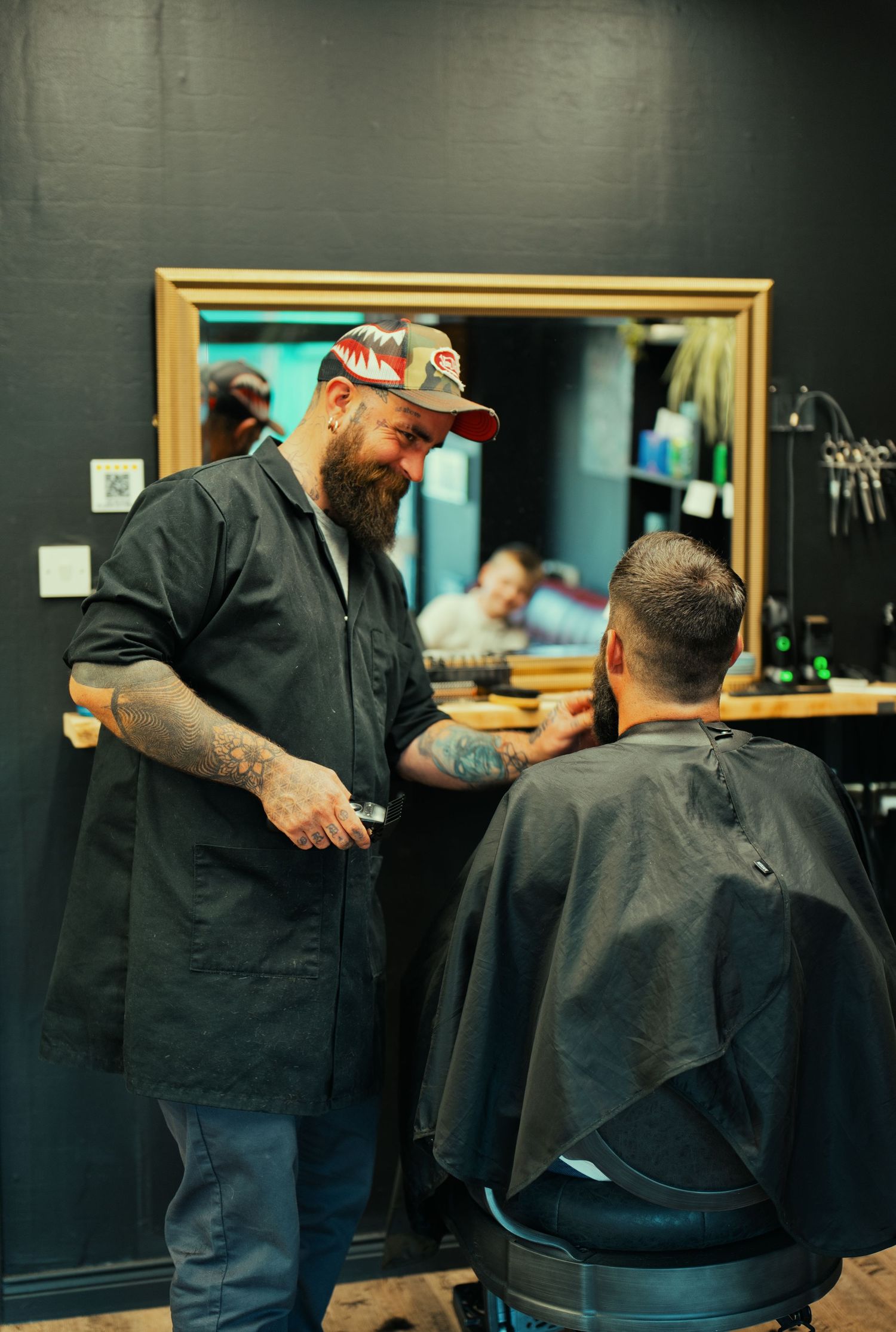 Tattooed barber in a cap consulting with a bearded client in a dark barbershop setting.
