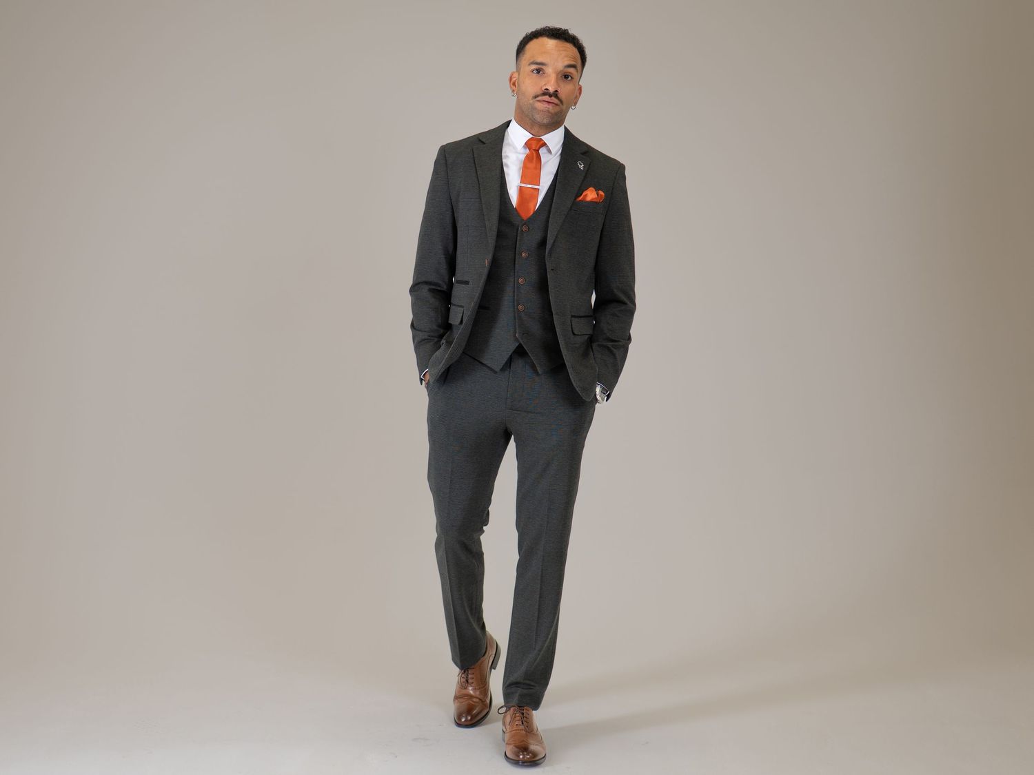Man in a charcoal three-piece suit with orange tie and pocket square walking confidently forward.