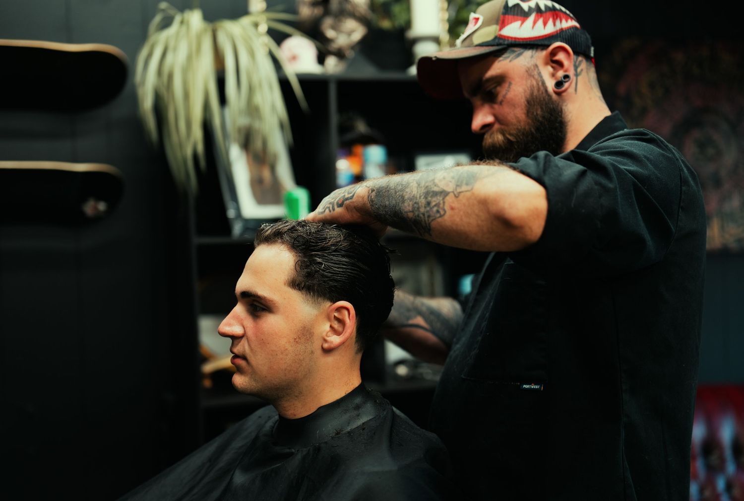 Tattooed barber styling a client's hair in a dark, edgy barbershop interior.