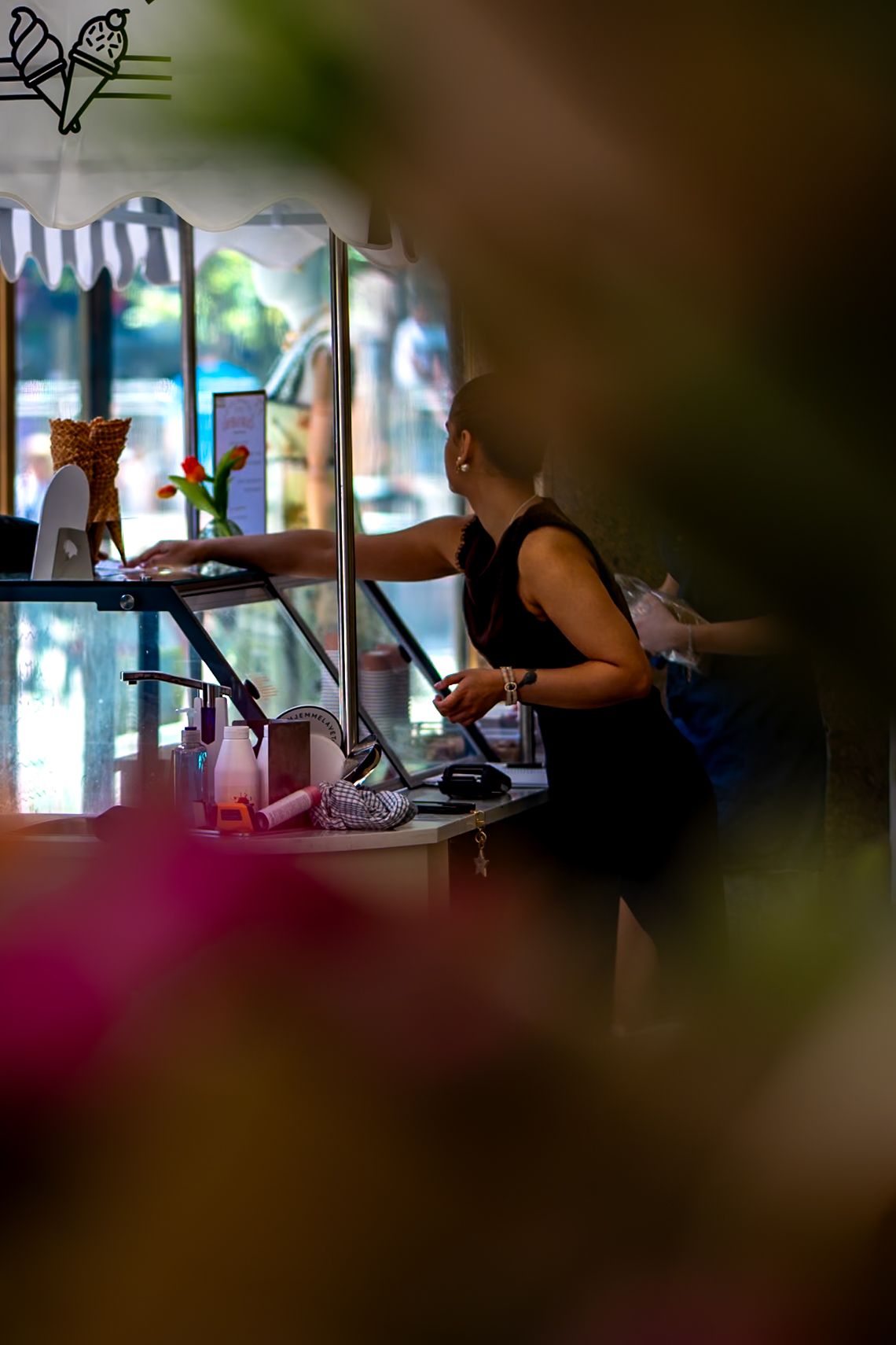 A bald person in black clothing works at an ice cream counter with a striped awning and floral decorations.