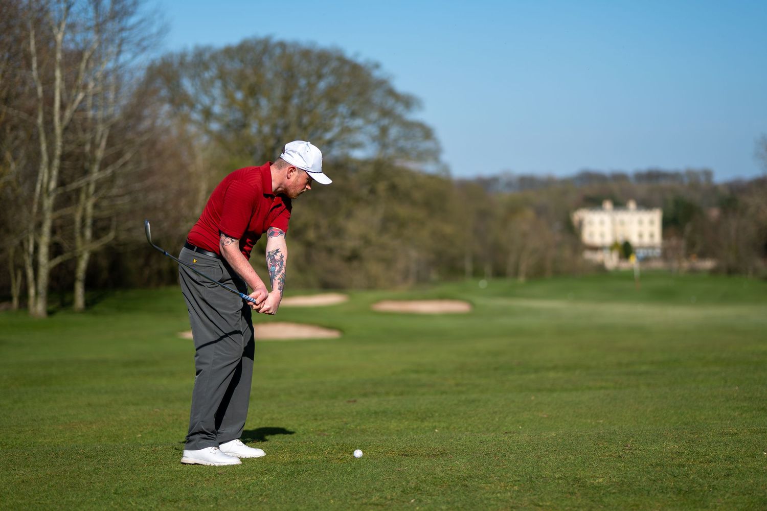 Golfer in red shirt and white cap preparing to take a shot on a lush green golf course.