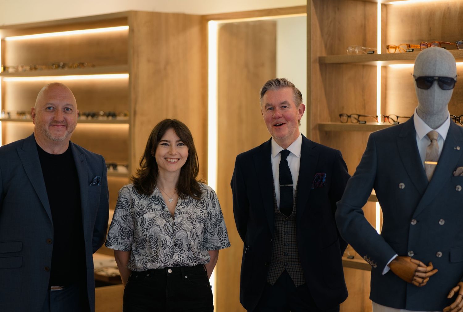 Three people smile beside a suited mannequin in a modern eyewear boutique with wooden display shelving.