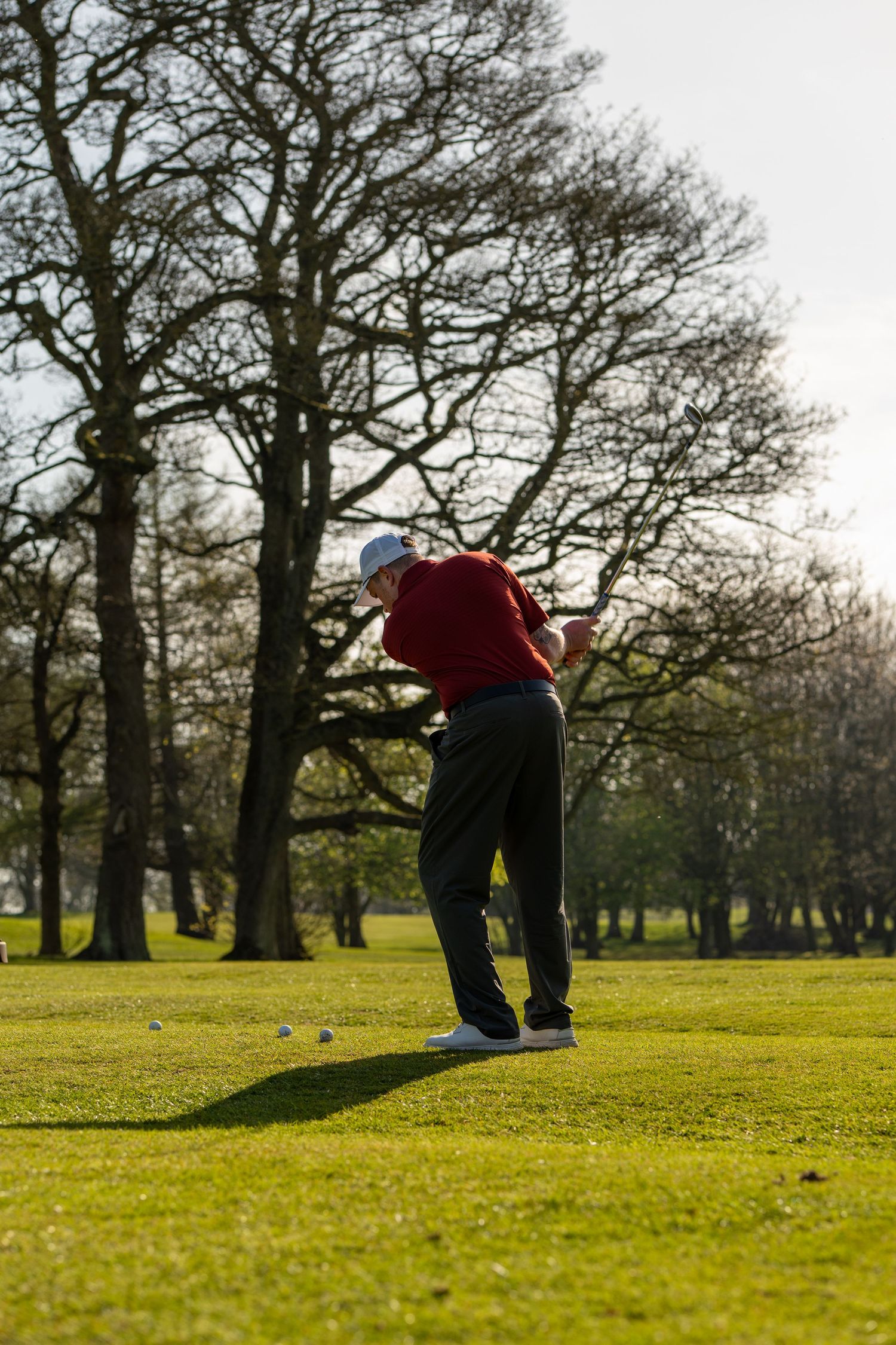 Golfer in red shirt and white cap completing golf swing on green fairway with bare winter trees behind.