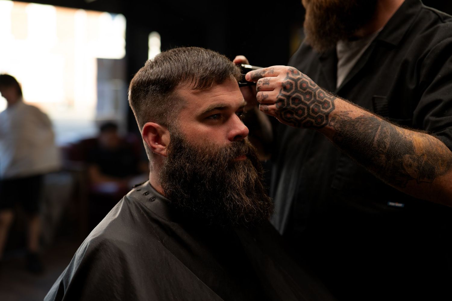 Tattooed barber trimming bearded man's hair with clippers in a professional barbershop.