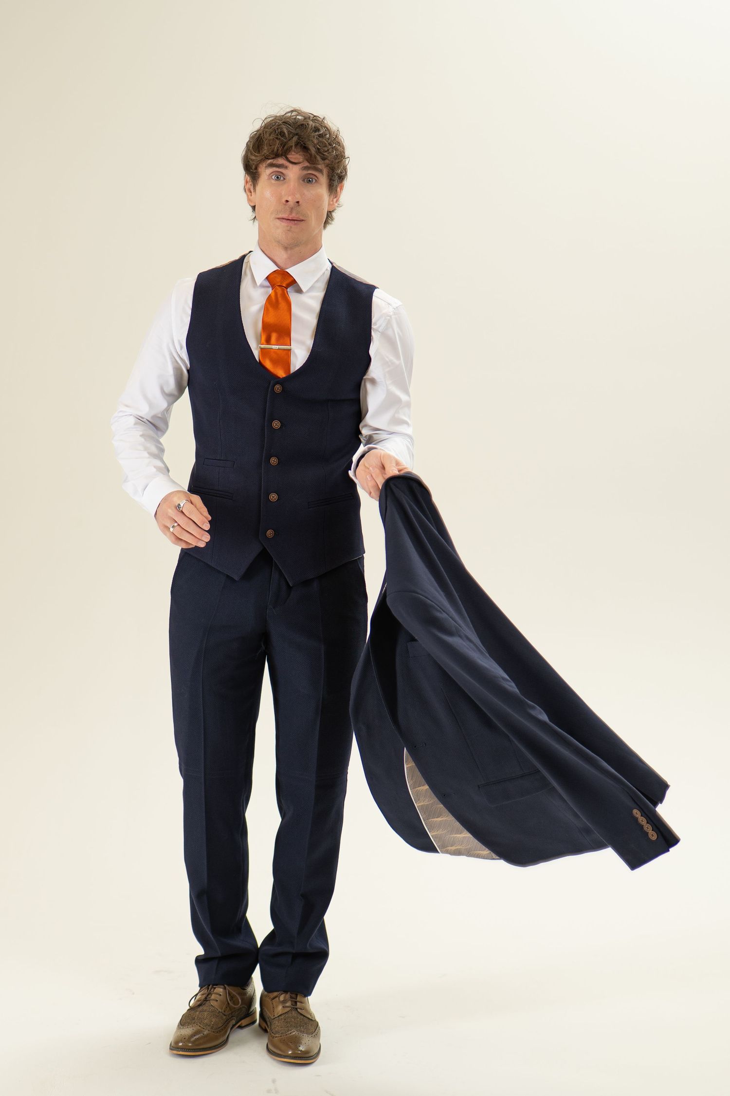 Man in navy waistcoat and orange tie holding a suit jacket to his side, studio shot.
