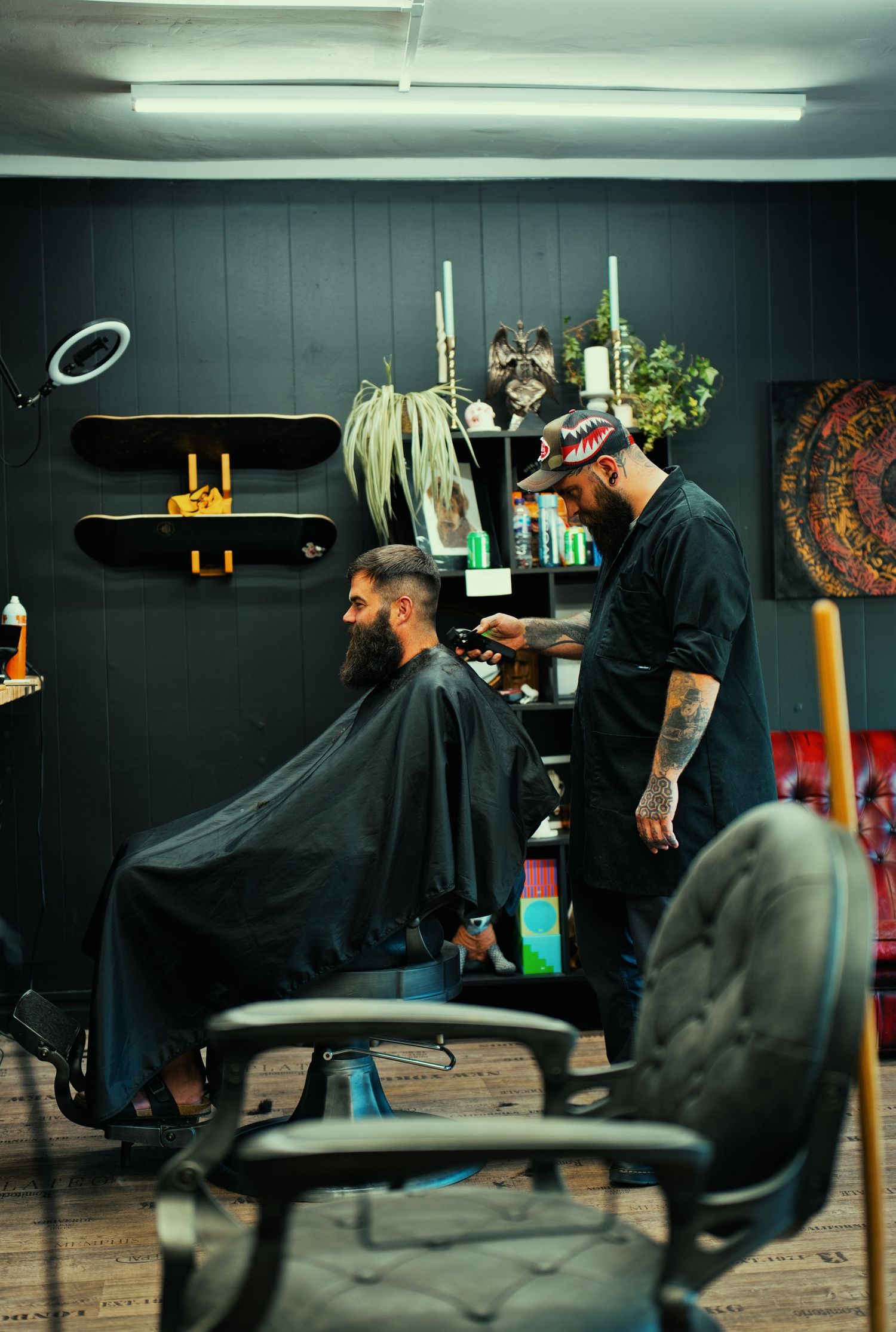 A tattooed barber cuts a bearded client's hair in a stylish dark-walled barbershop.
