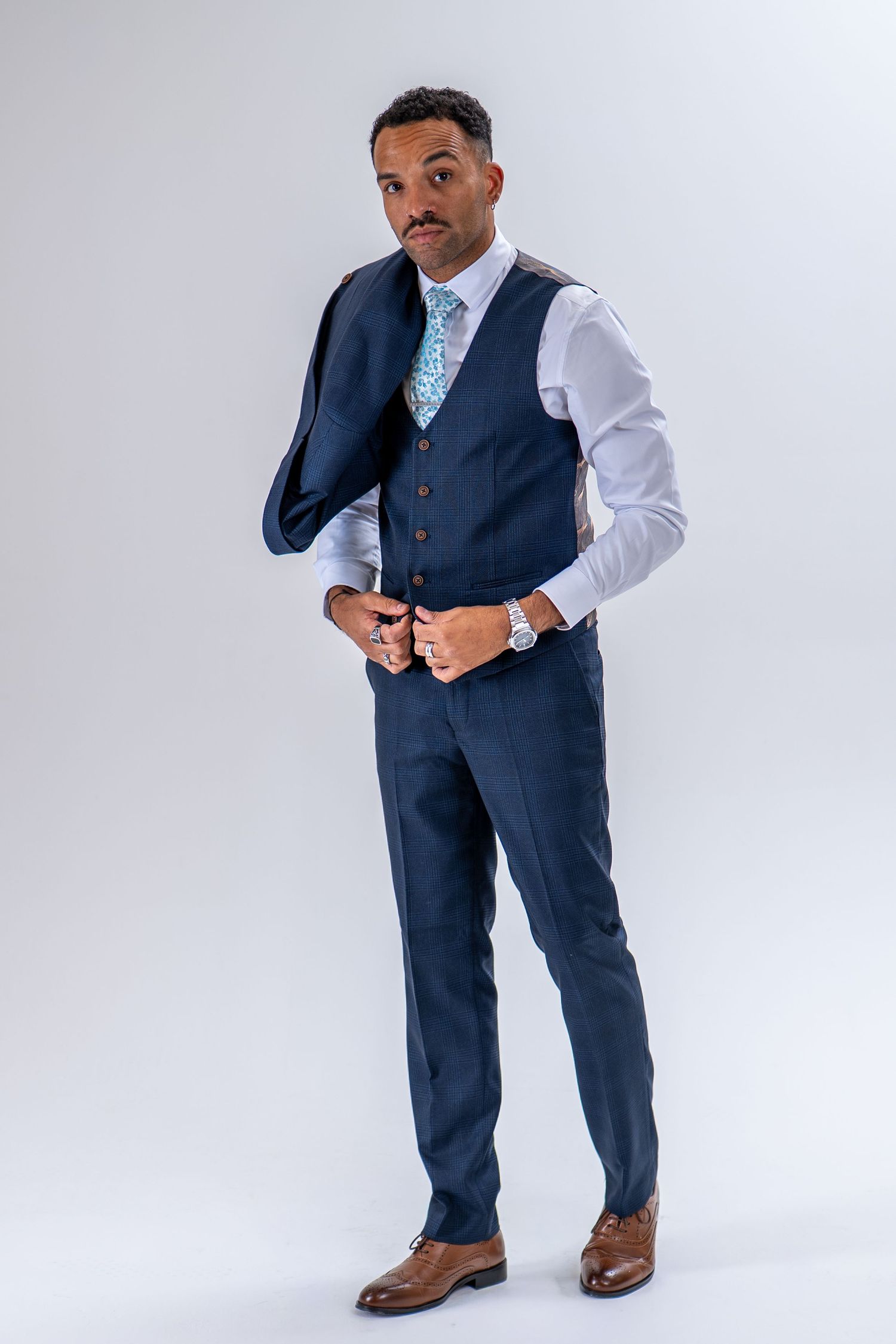 Man in navy blue waistcoat and trousers adjusting jacket, wearing silver watch and blue floral tie.