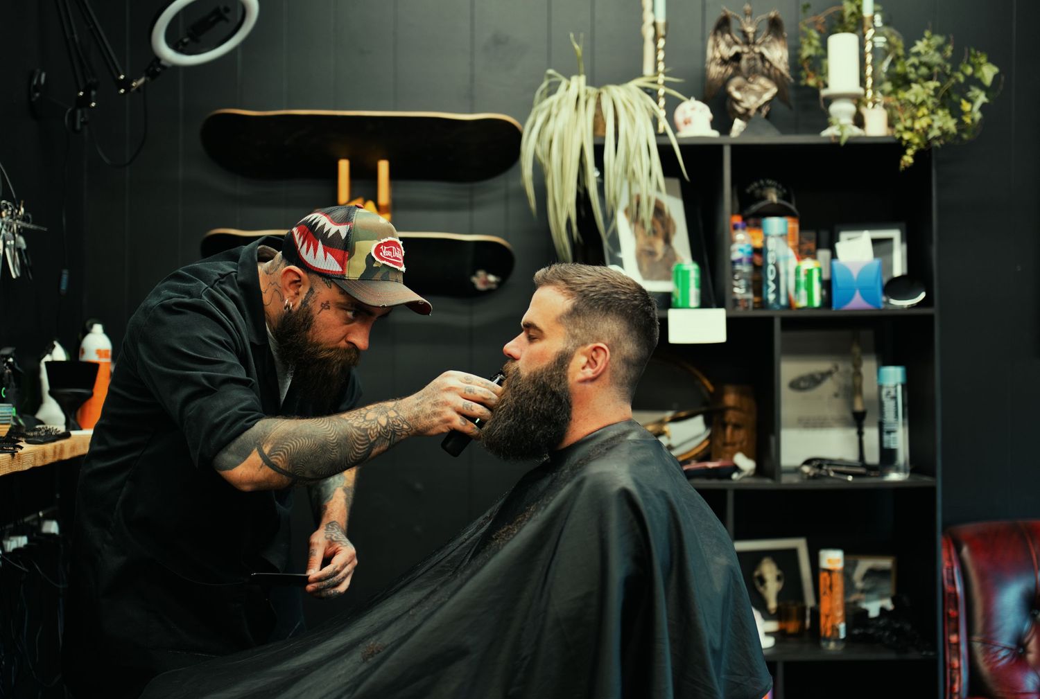 Tattooed barber trimming beard of client in stylish dark-themed barbershop.