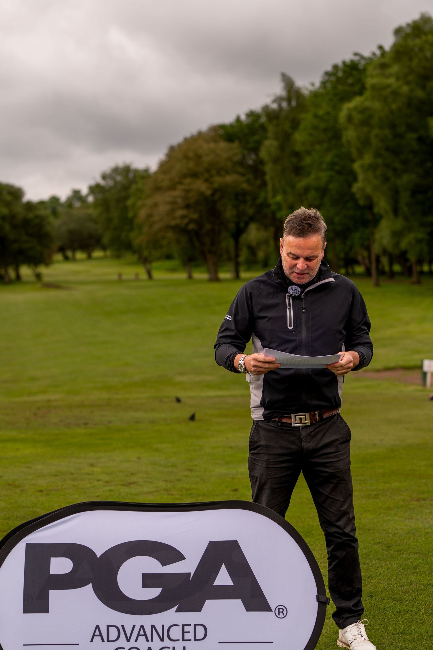 PGA Advanced Coach standing on golf course reading papers beside a PGA branded banner.