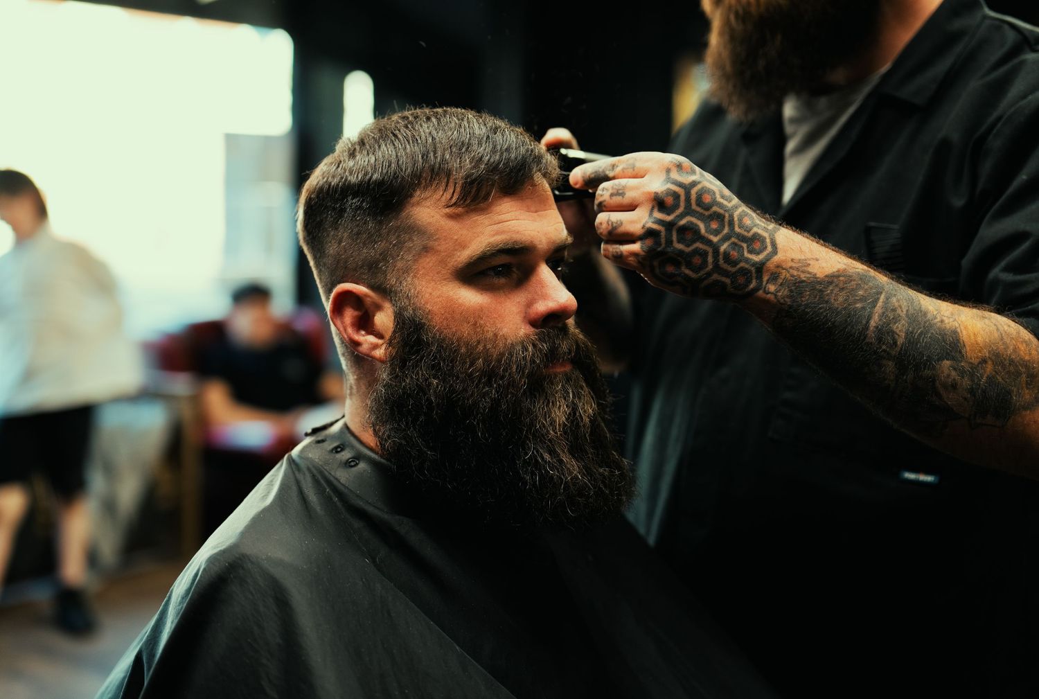 A tattooed barber uses clippers to style a bearded man's hair in a busy barbershop.