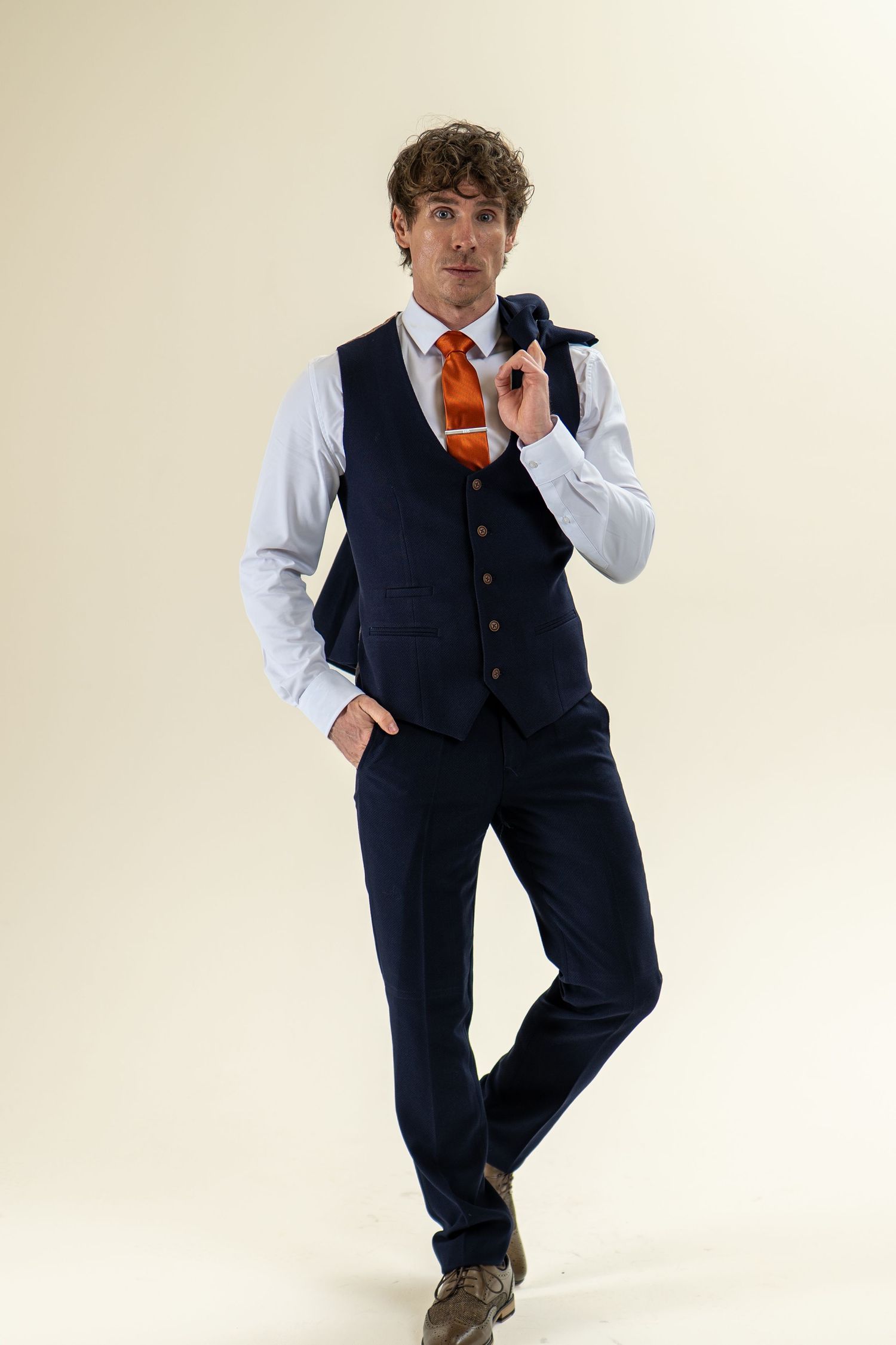 Man in navy waistcoat and orange tie holding jacket over shoulder, studio portrait.