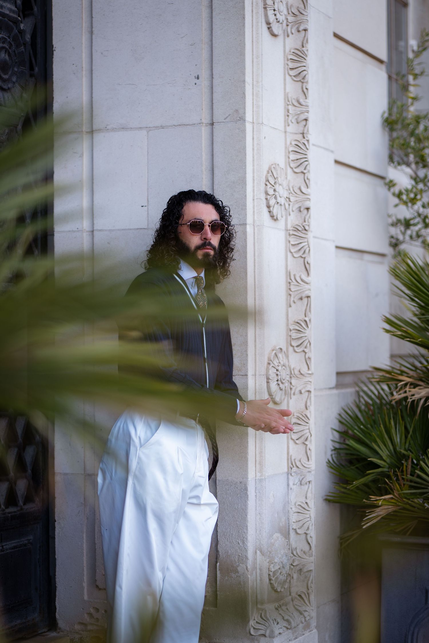Curly-haired man in green jacket and white trousers beside decorative stone column.