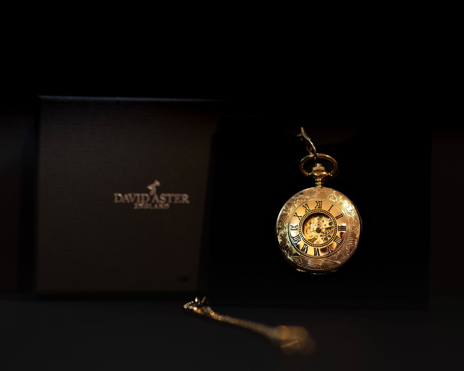 Luxury gold pocket watch displayed beside David Aster Jeweller black gift box on dark background.