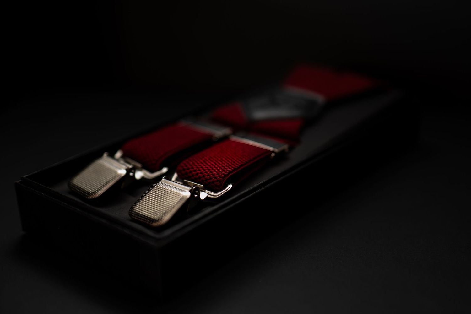 Close-up of red suspenders with silver metal clips neatly arranged in black presentation box.