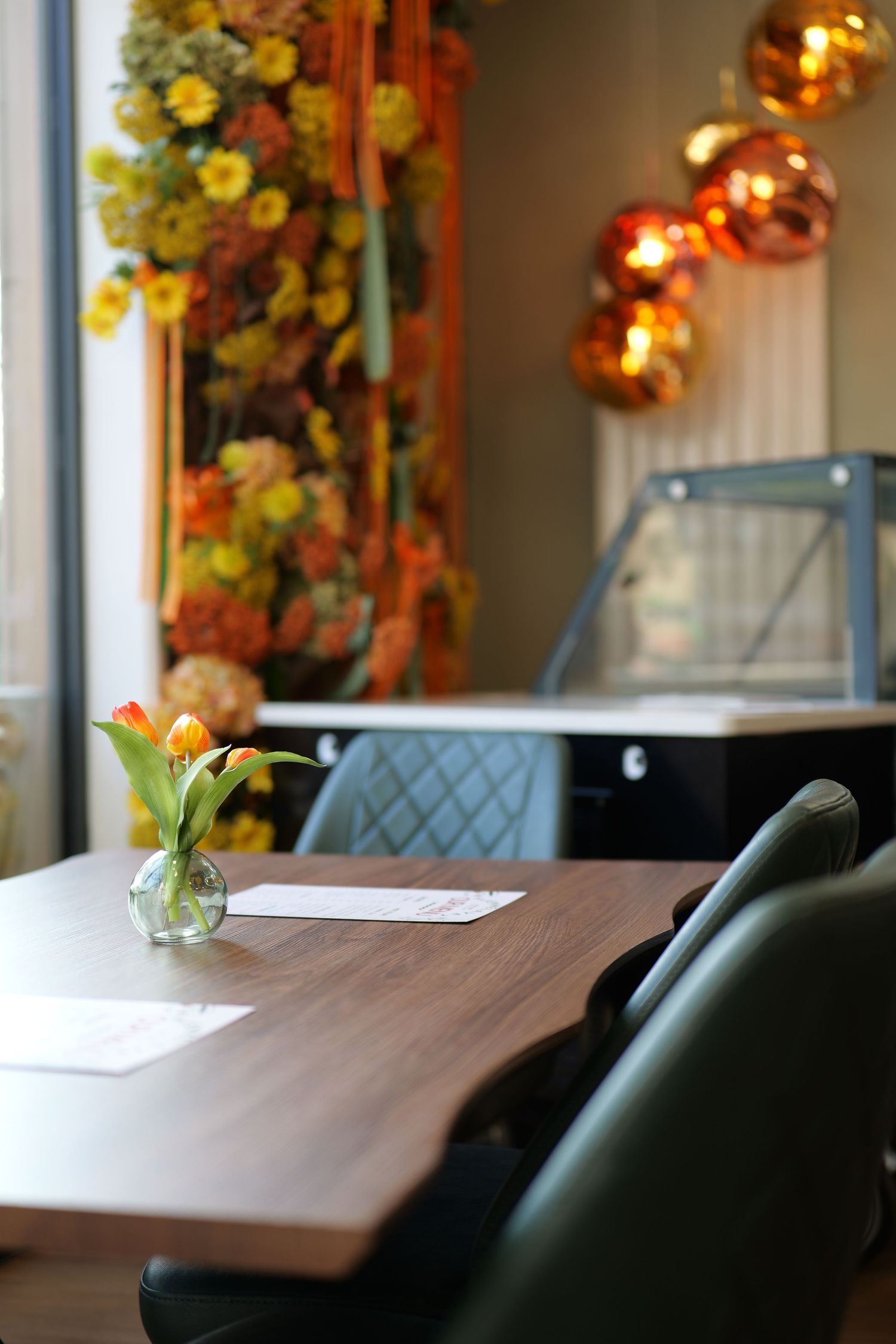Cozy cafe interior with tulip centerpiece, green chairs, golden pendant lights, and floral wall decor.