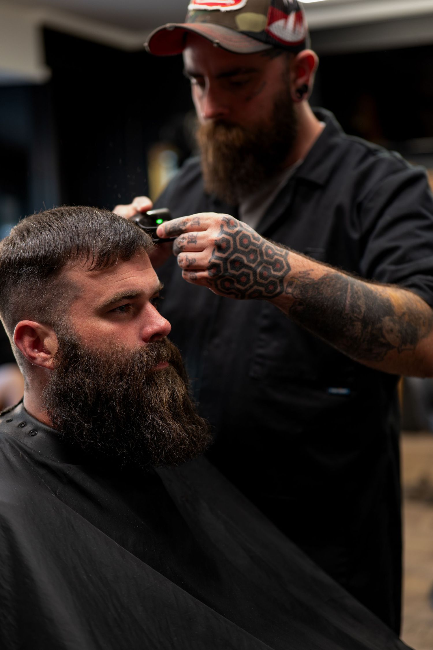 Tattooed barber in cap trimming bearded clients hair in professional barbershop.