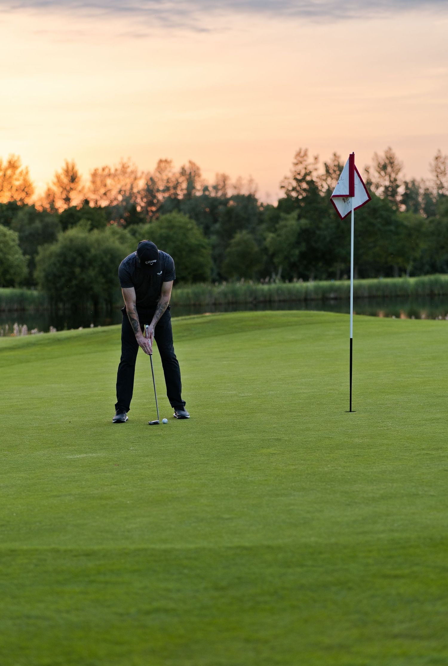 A golfer putts on a green at sunset with a red and white flag pin visible on the course.