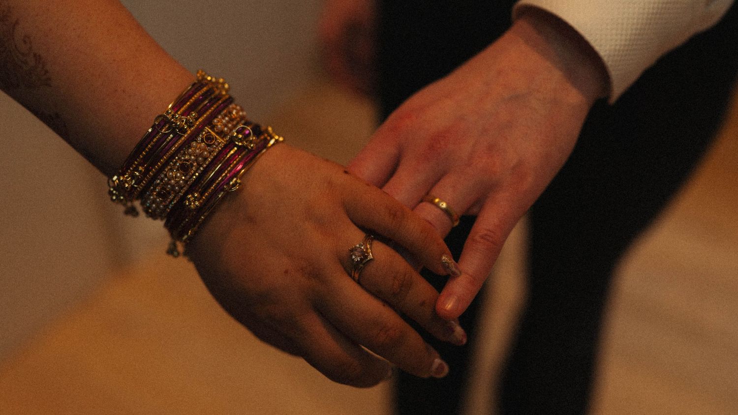 Close-up of two hands holding, one adorned with gold bangles and a ring, the other wearing a simple gold band.