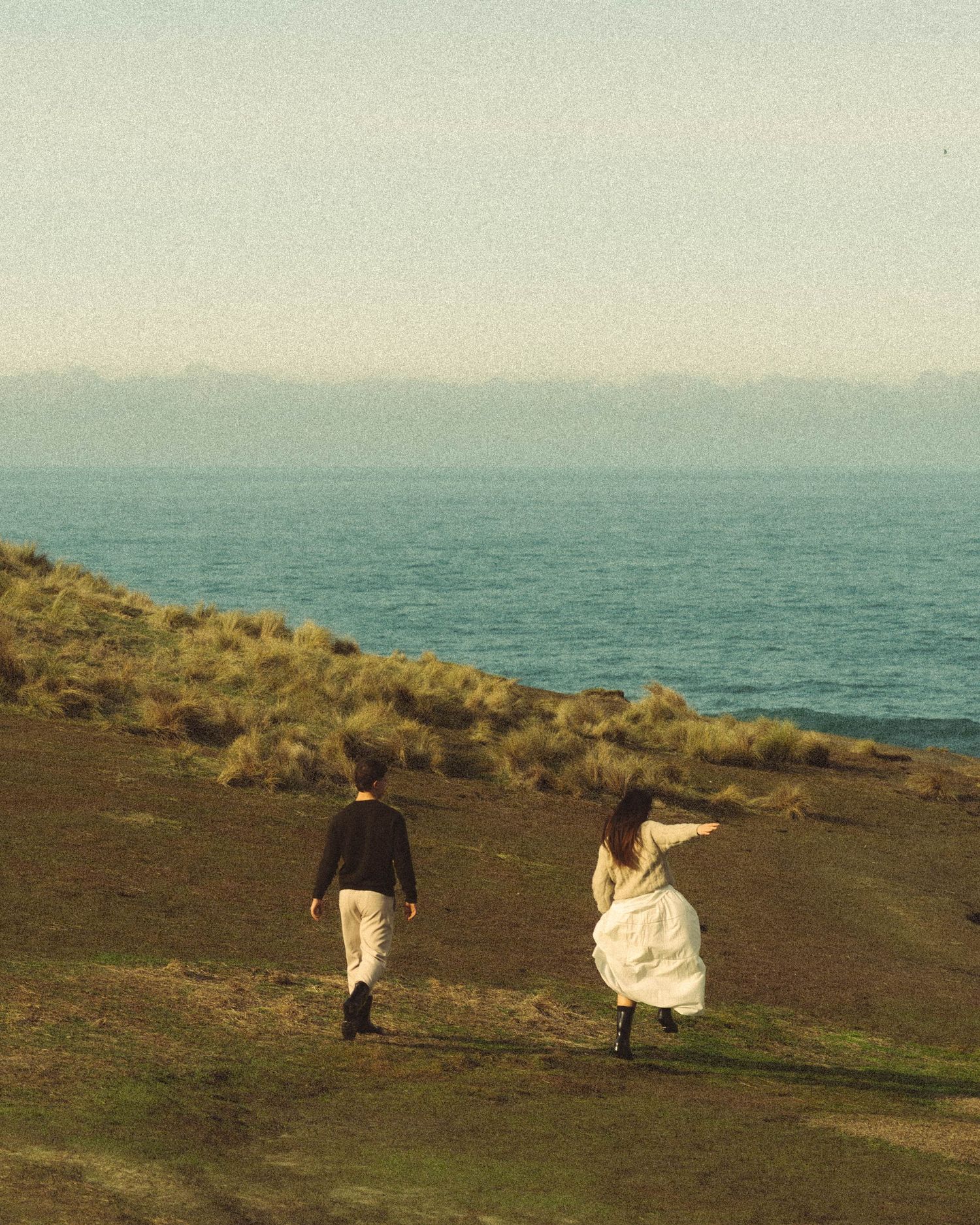 Couple strolling toward turquoise ocean horizon across grassy coastal headland.