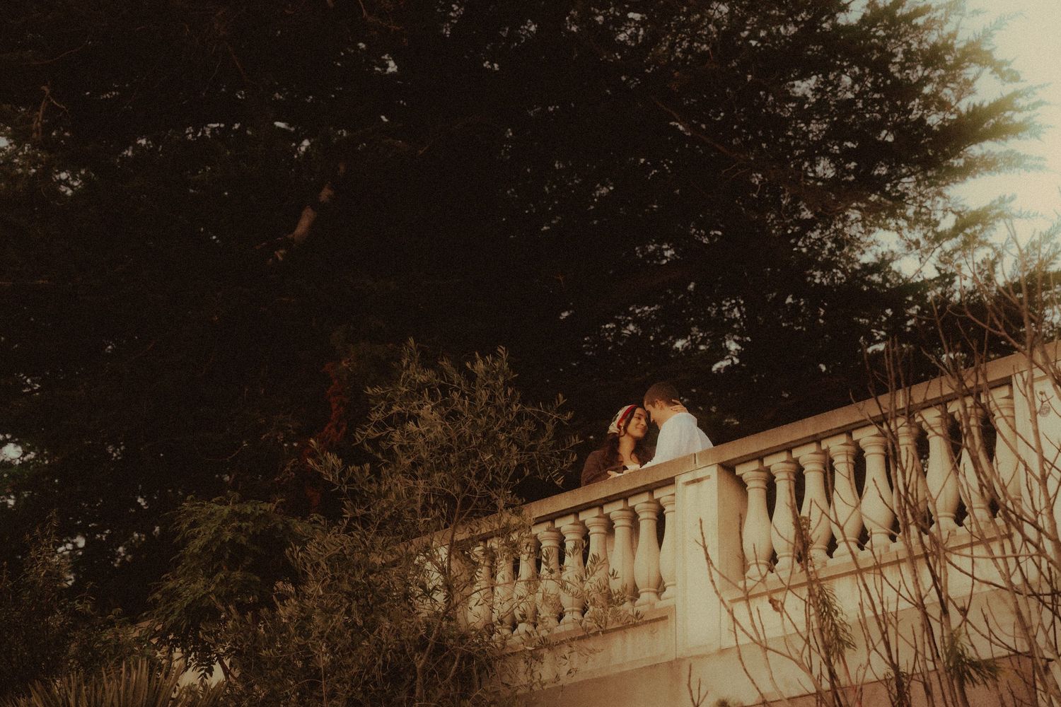 Wedding couple sharing a kiss on an ornate stone balcony surrounded by lush trees.