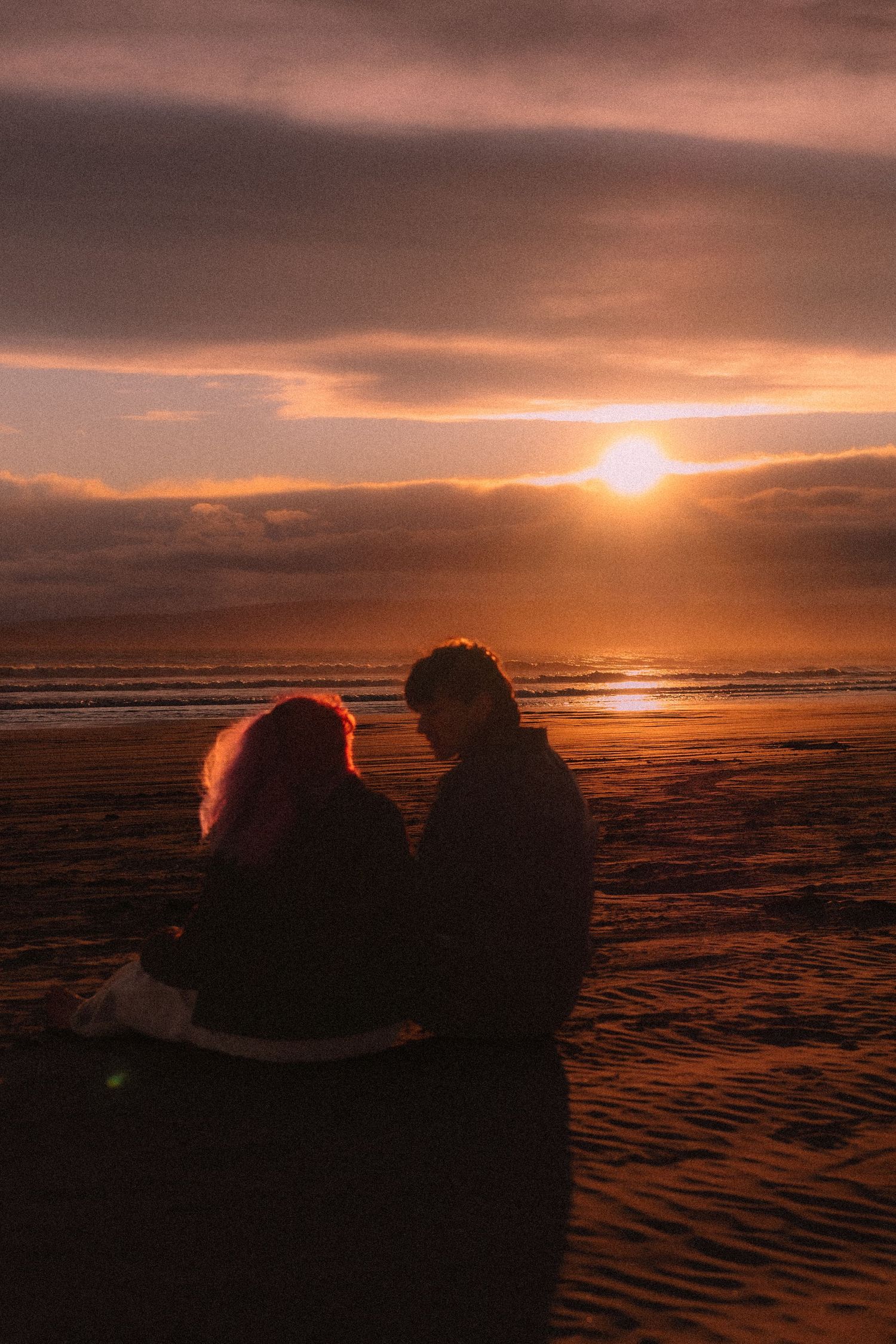 Couple silhouetted against a stunning golden sunset on a sandy beach, sitting together romantically.
