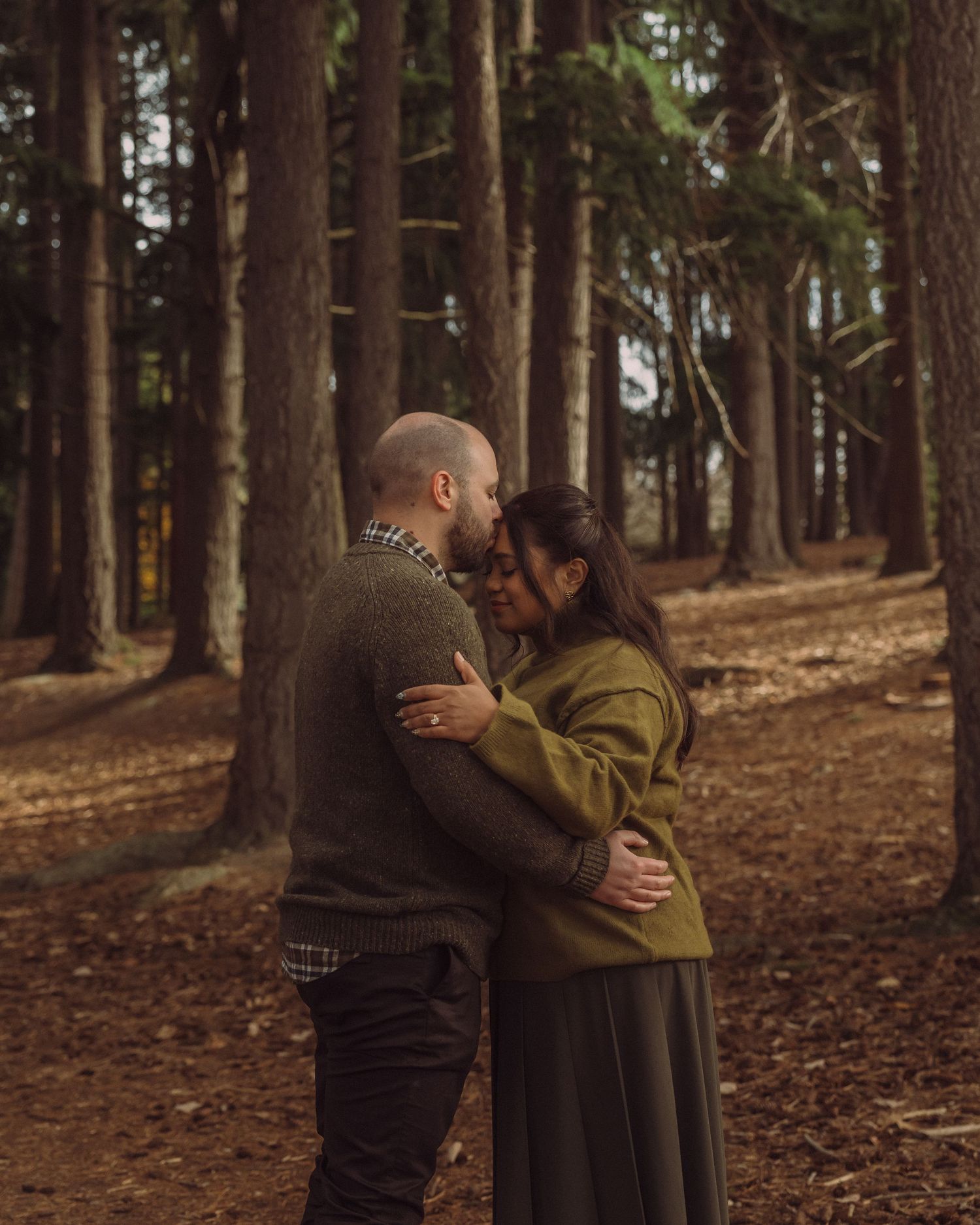 Couple embracing romantically among tall pine trees in an autumn forest setting.