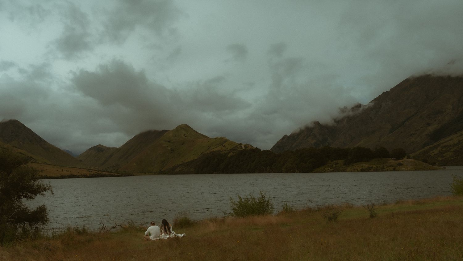 Couple sitting by tranquil lake with dramatic cloudy mountain scenery. Located in Queenstown, New Zealand