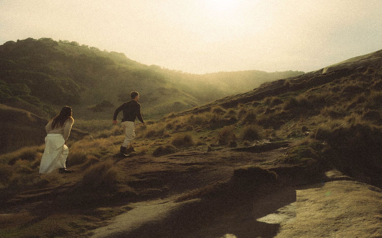 Couple walking up rocky hillside trail bathed in warm golden sunset light.