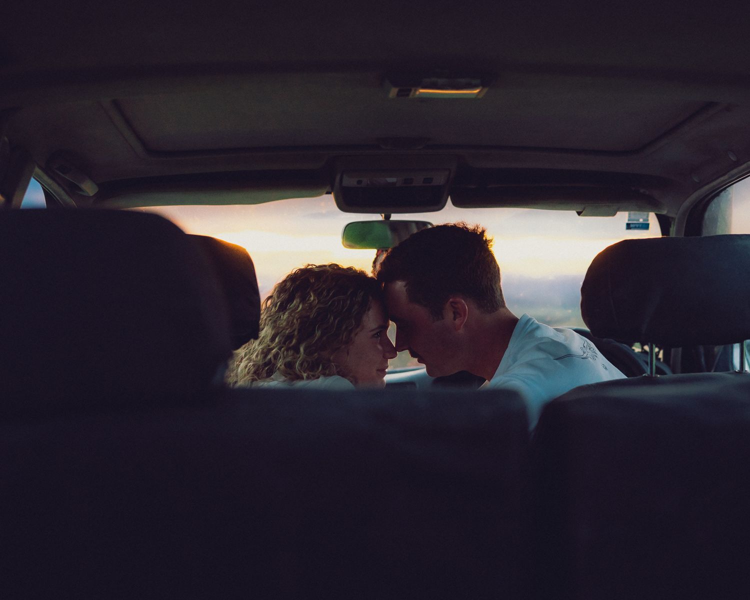 Romantic couple leaning close together inside a car at sunset, sharing an intimate moment.