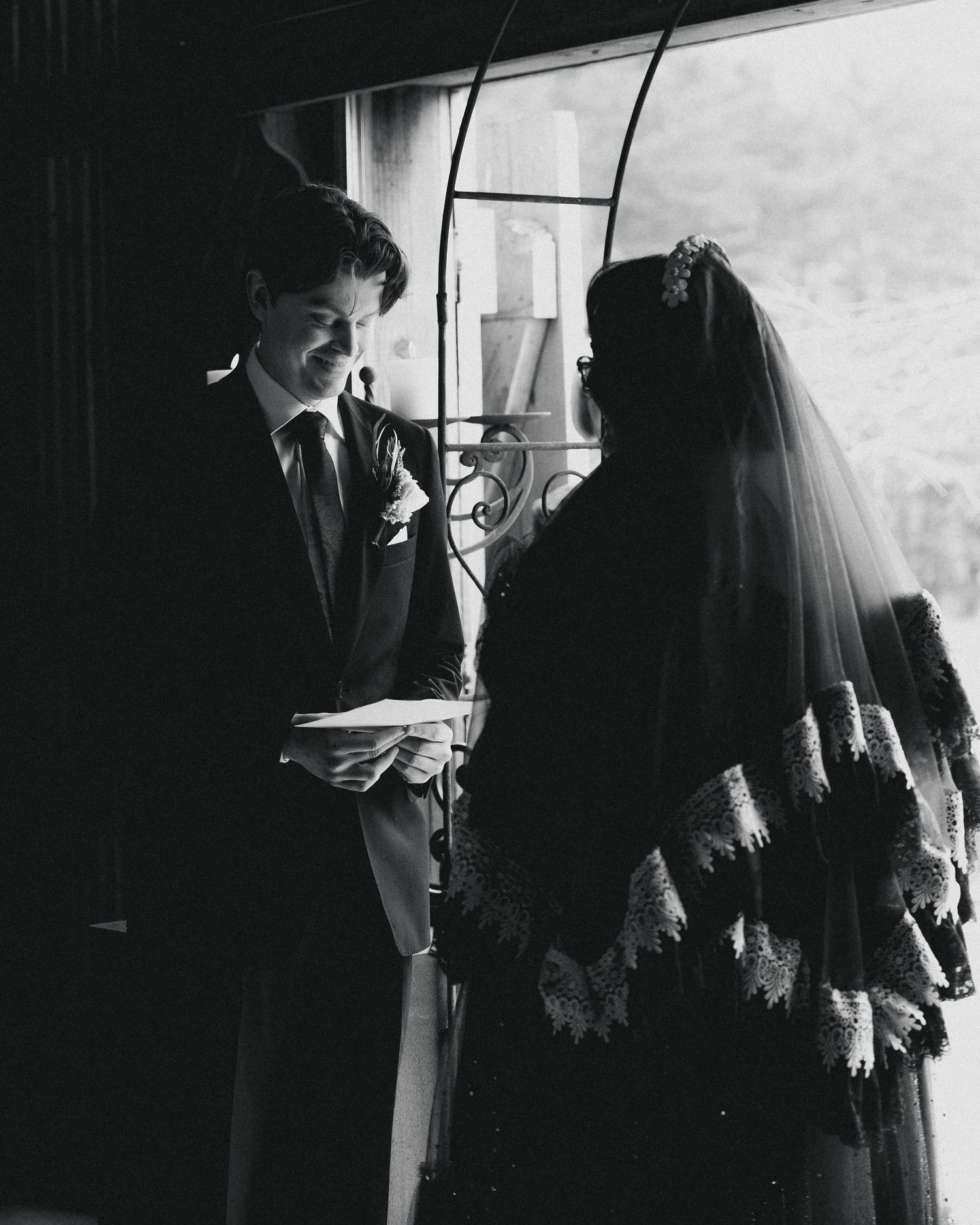 Black and white photo of a groom reading vows to a veiled bride in a dark wedding gown at an elegant ceremony.