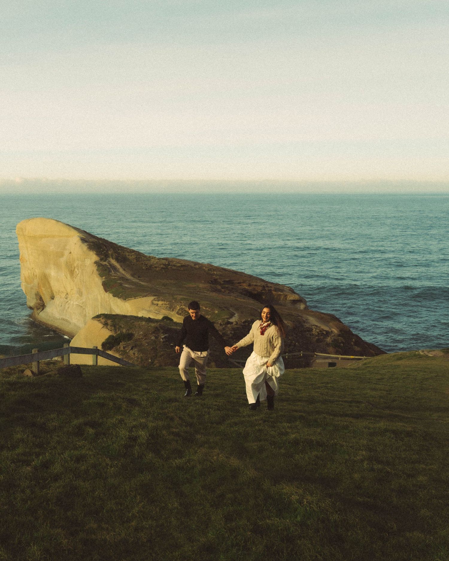 Couple holding hands running past large coastal rock with ocean backdrop. Located in Tunnel Beach, Dunedin