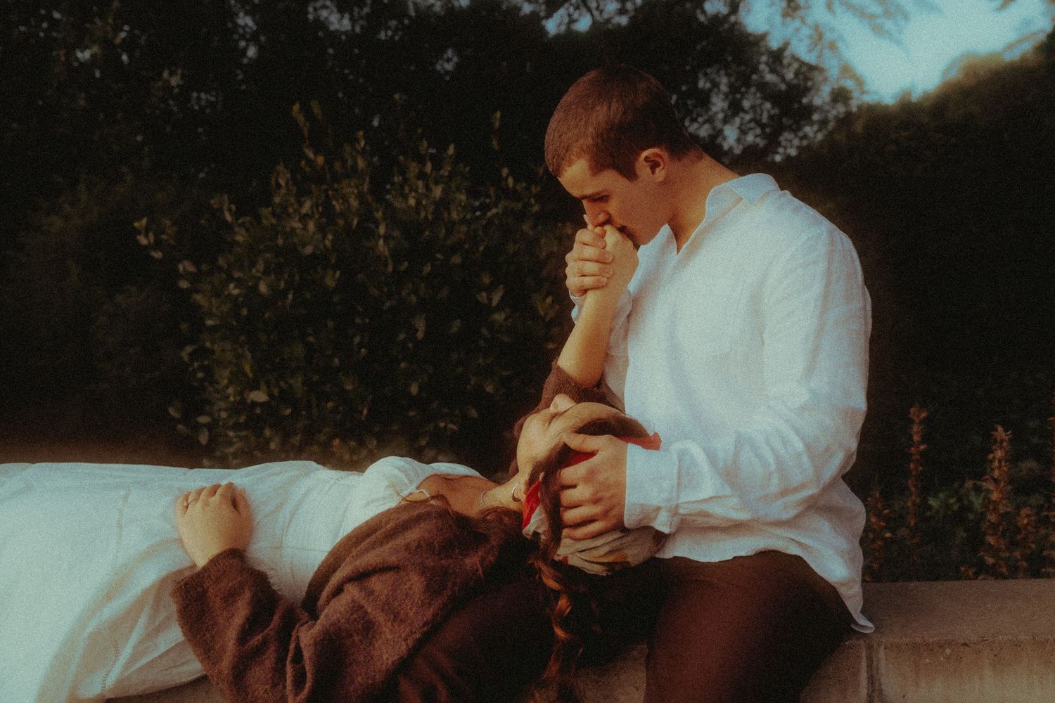 Man tenderly kissing womans hand as she rests outdoors in soft golden light.