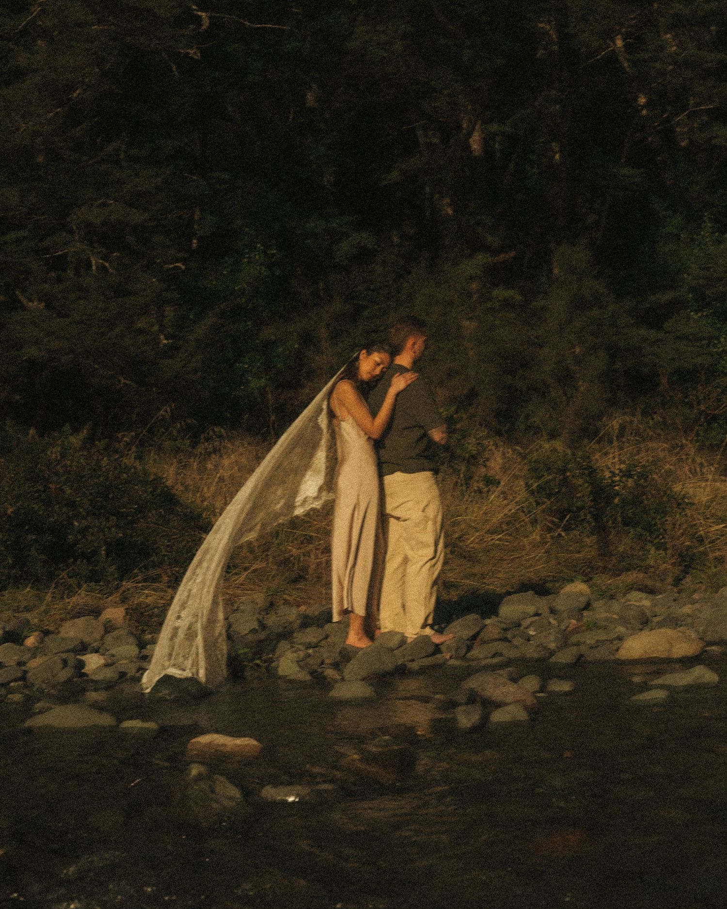Couple sharing an intimate embrace by a rocky riverside surrounded by dark forest trees.