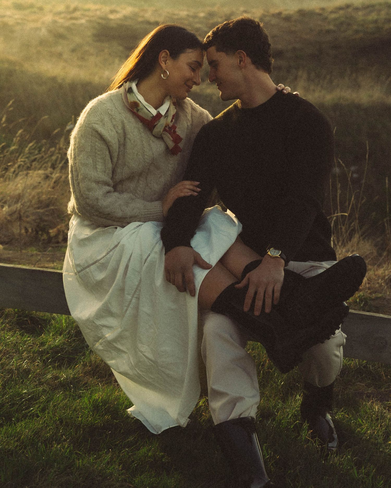 Intimate couple leaning close together seated on wooden fence at golden hour.