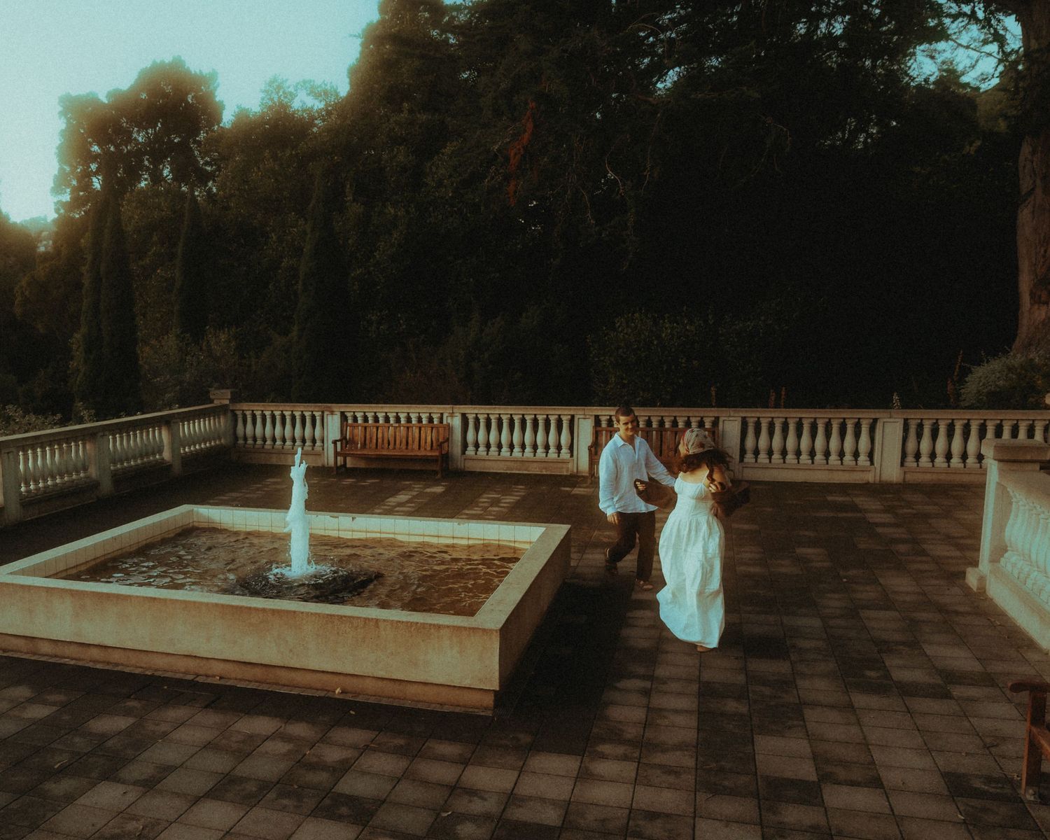 Couple in formal attire standing by a garden fountain on a stone terrace surrounded by lush trees.