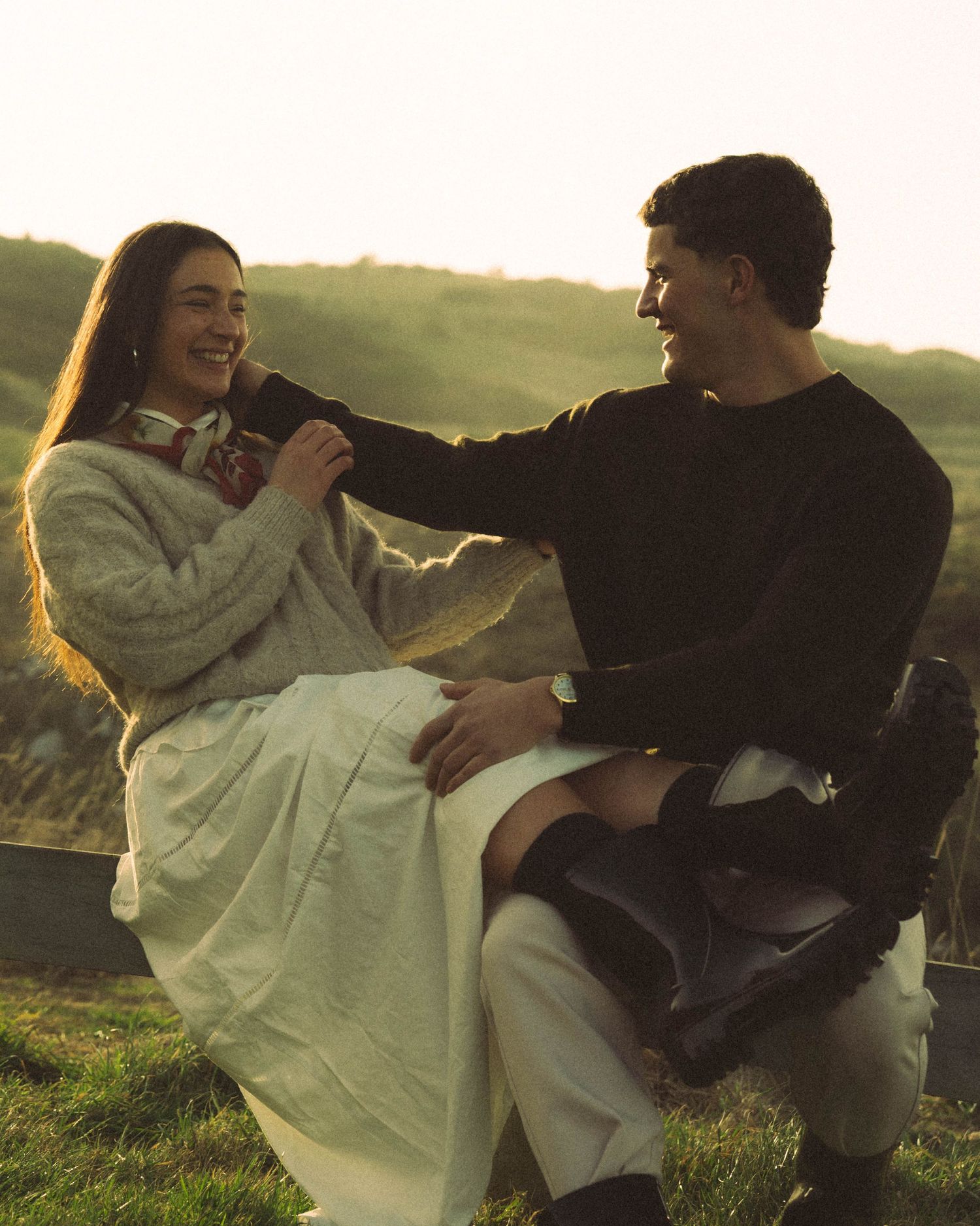 Couple laughing together on a wooden fence in a sunlit green hillside, woman in grey sweater and white skirt. Located in Dunedin, New Zealan