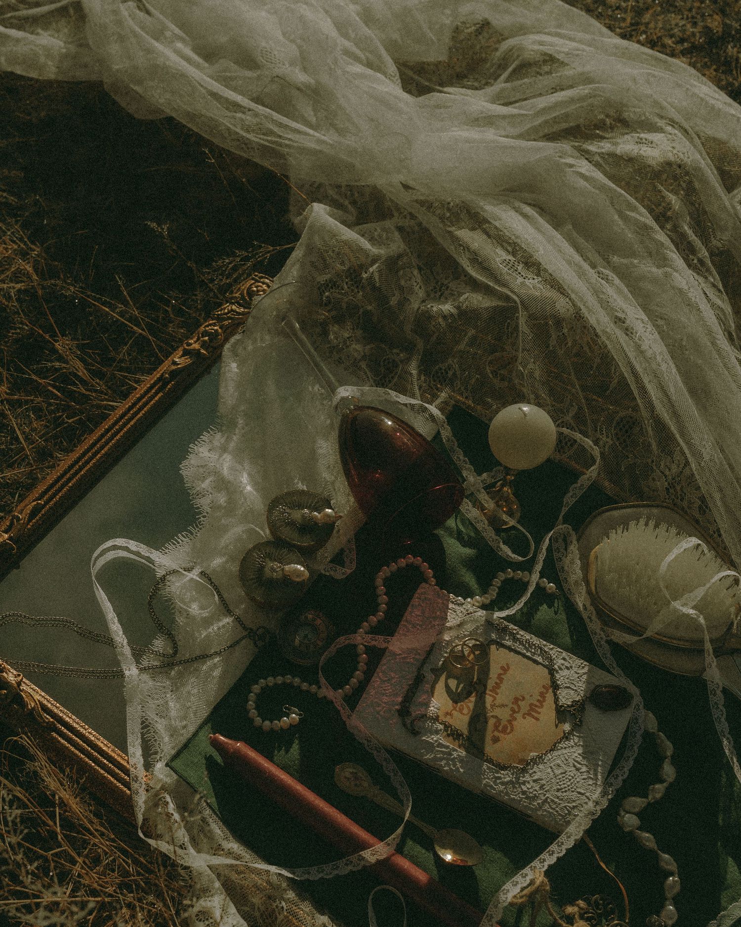 Vintage flat lay with pearls, red gem, lace fabric, antique books, and jewelry arranged on grass outdoors.